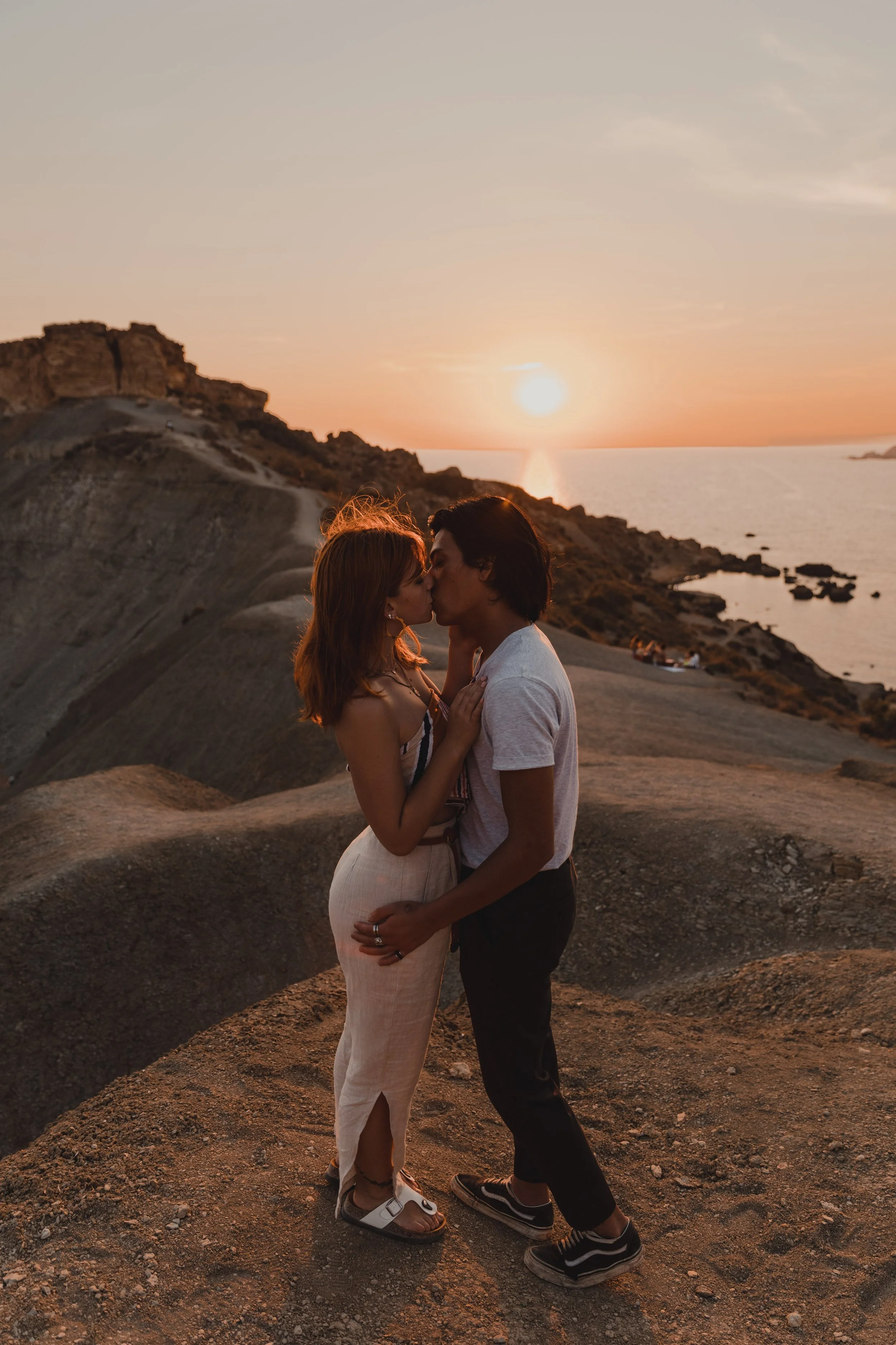 Couple kissing on a rocky cliff during sunset overlooking the sea.