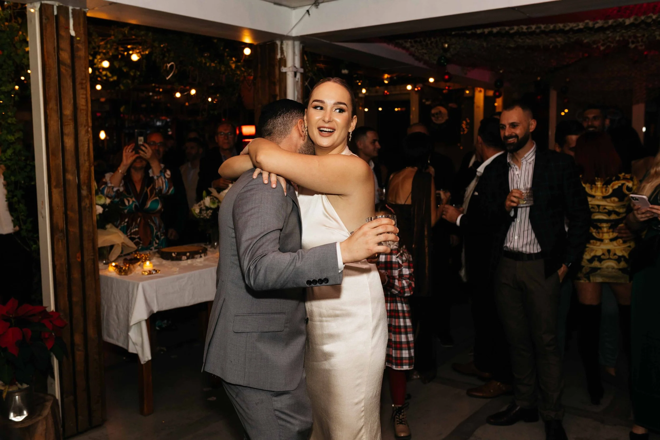 A happy couple embracing at a lively indoor party, surrounded by guests, with festive decorations and dim lighting.