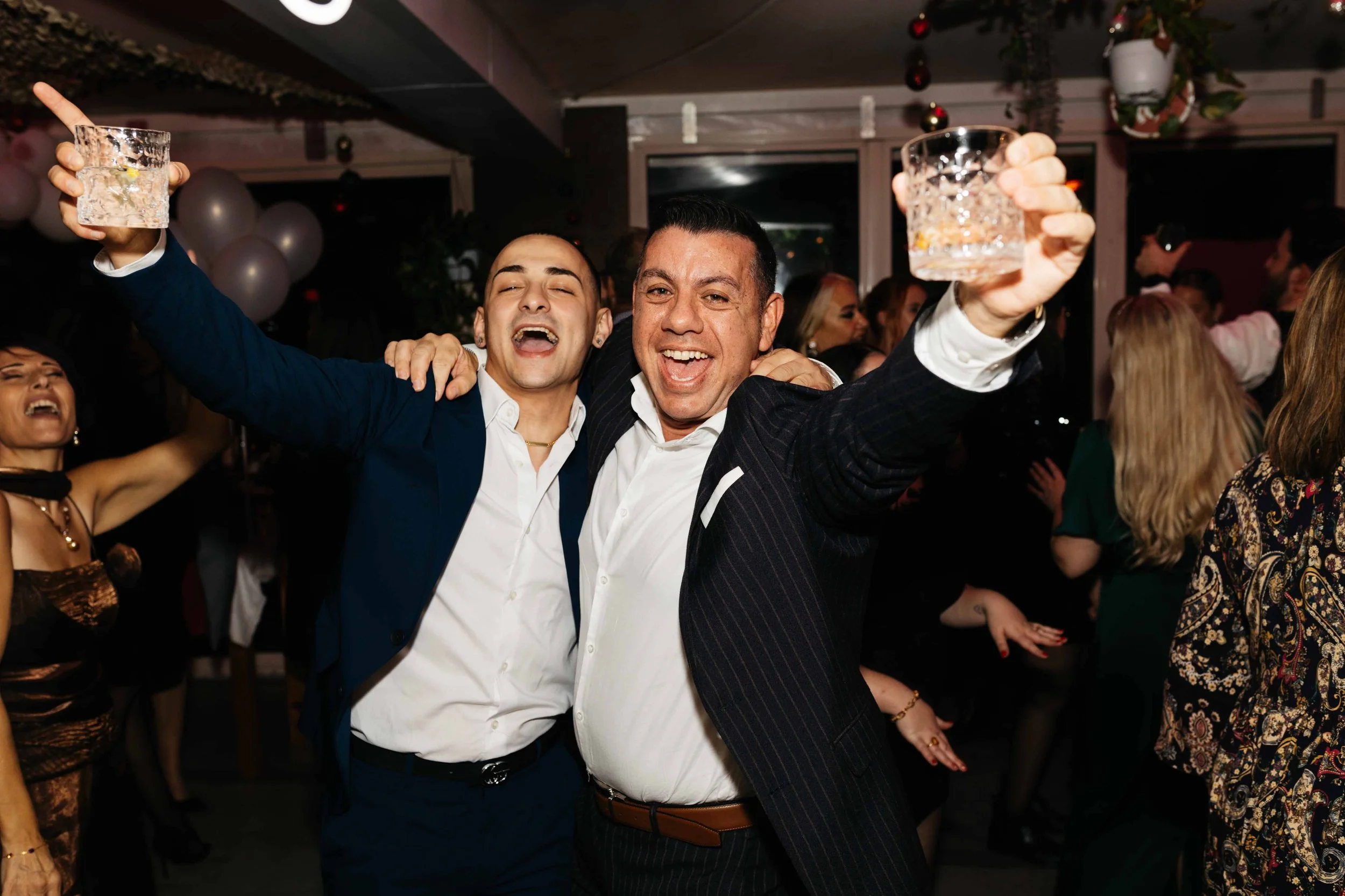 Two men in suits celebrating at a party, holding drinks, with people dancing in the background.