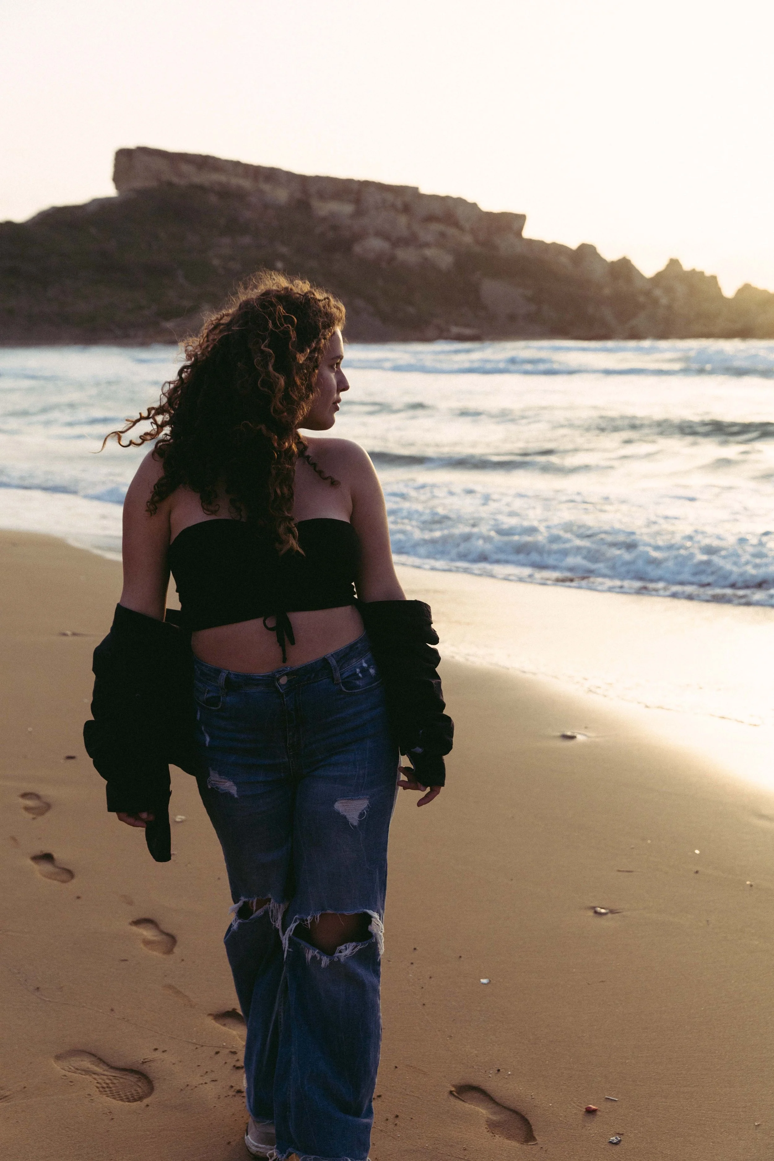 Woman with curly hair walking on a sandy beach near ocean waves during sunset.