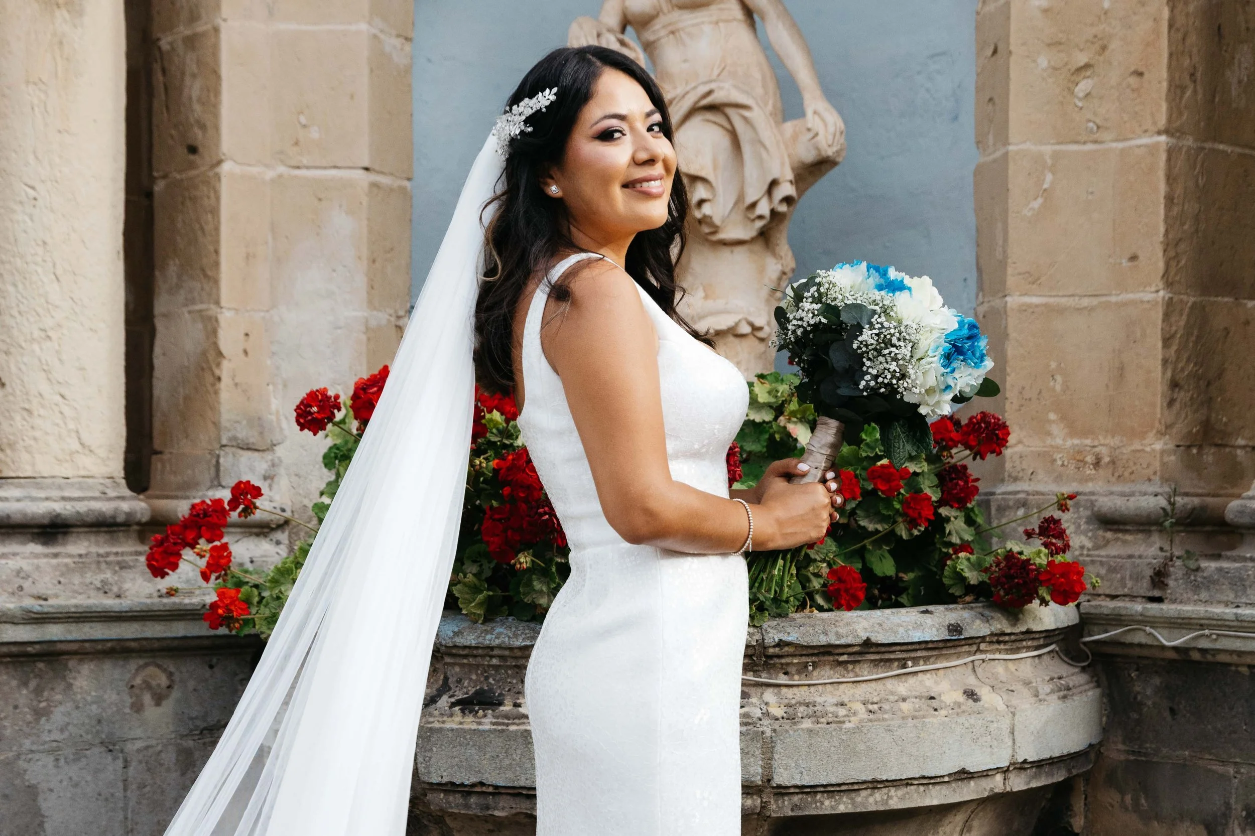 Bride in white dress holding bouquet in front of stone wall with red flowers