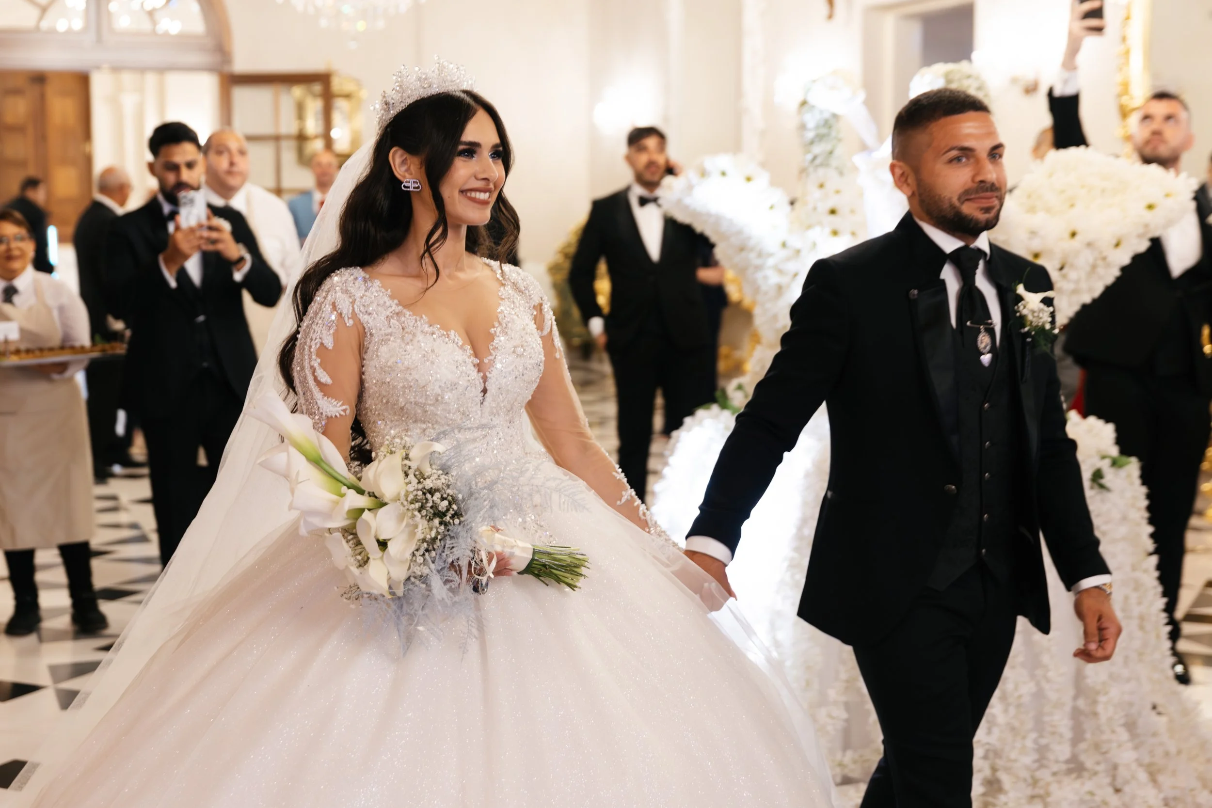 Happy bride and groom walking hand in hand during wedding ceremony, surrounded by guests in formal attire, bride wearing a white gown and tiara, groom in a dark suit.