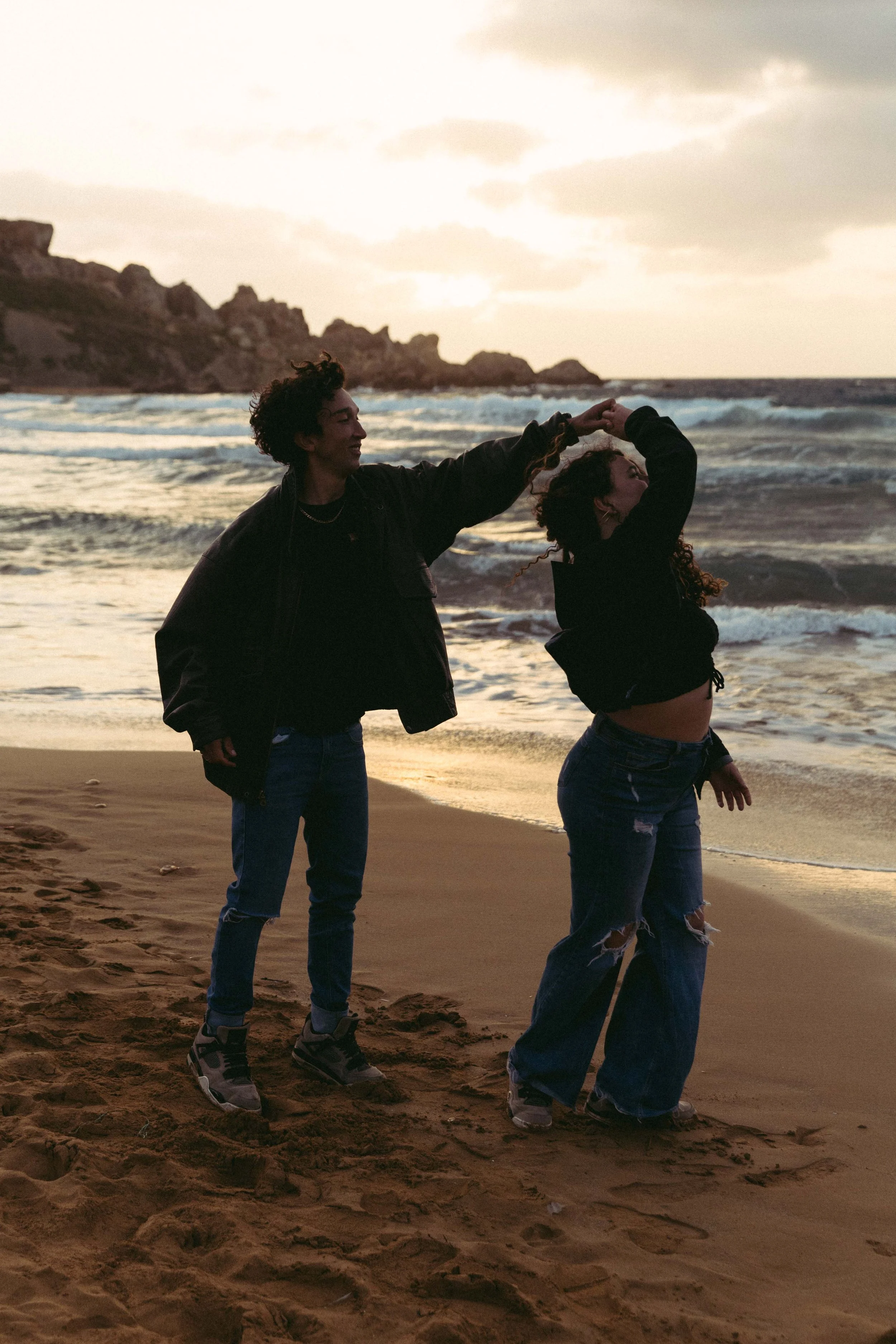 Two people dancing on a beach as the sun sets, with ocean waves in the background.