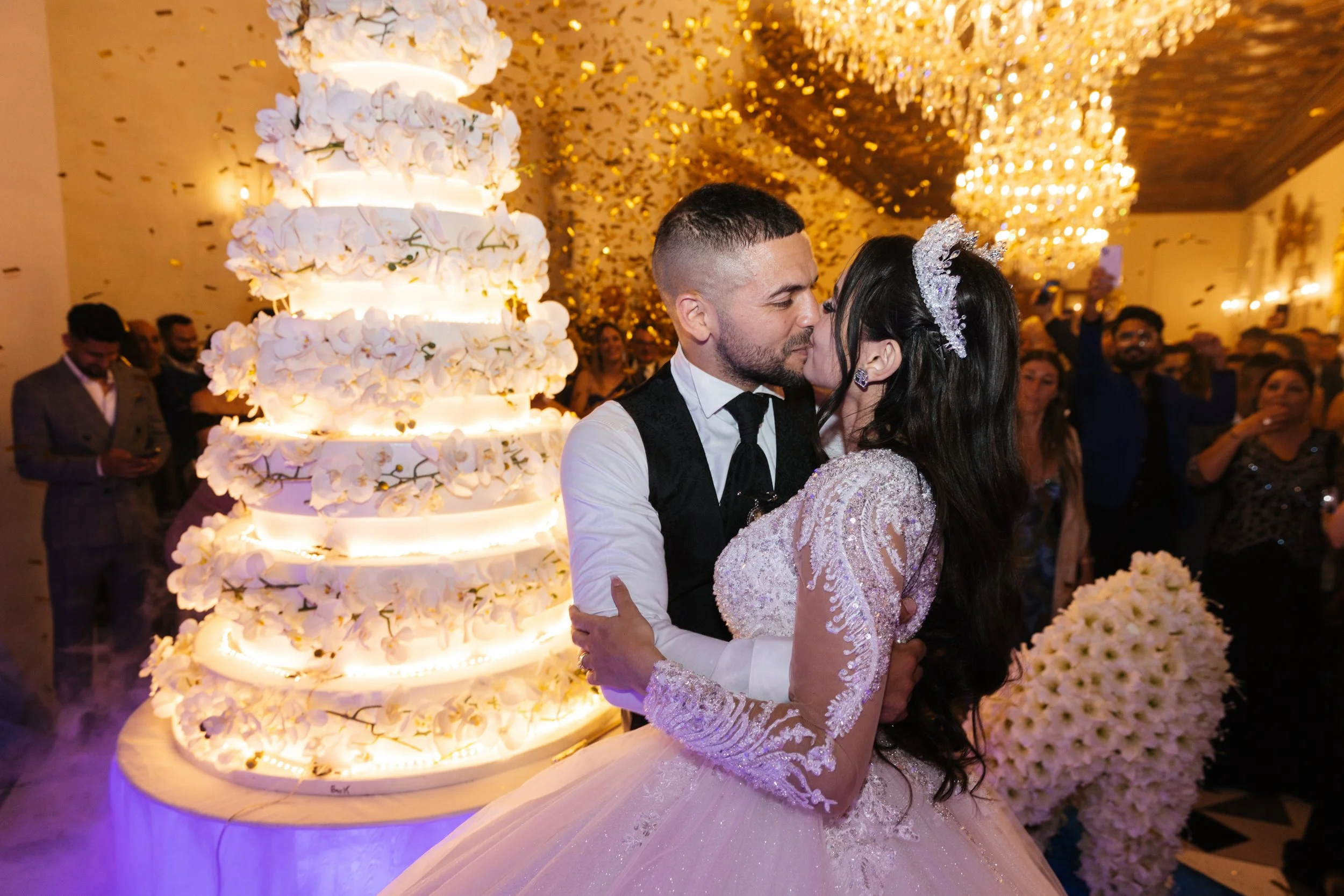 Wedding couple kissing near large white cake with floral decorations, surrounded by confetti and chandeliers.