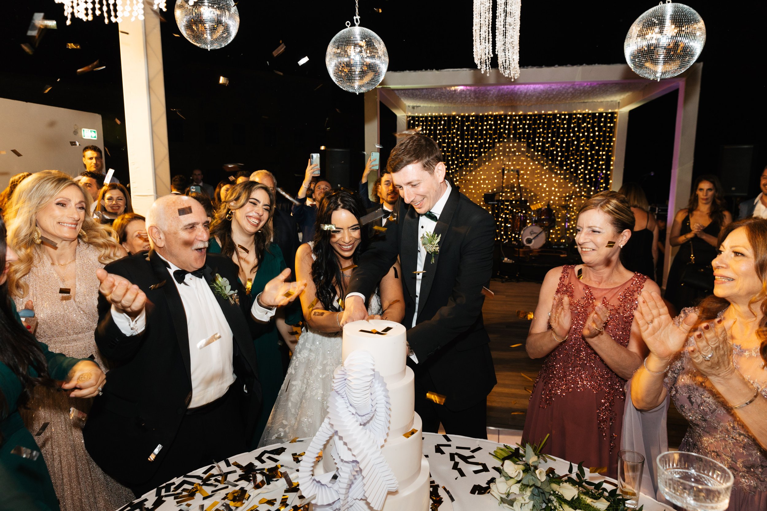 Bride and groom cutting wedding cake at a celebration with guests clapping and confetti falling, surrounded by elegant decorations and string lights.