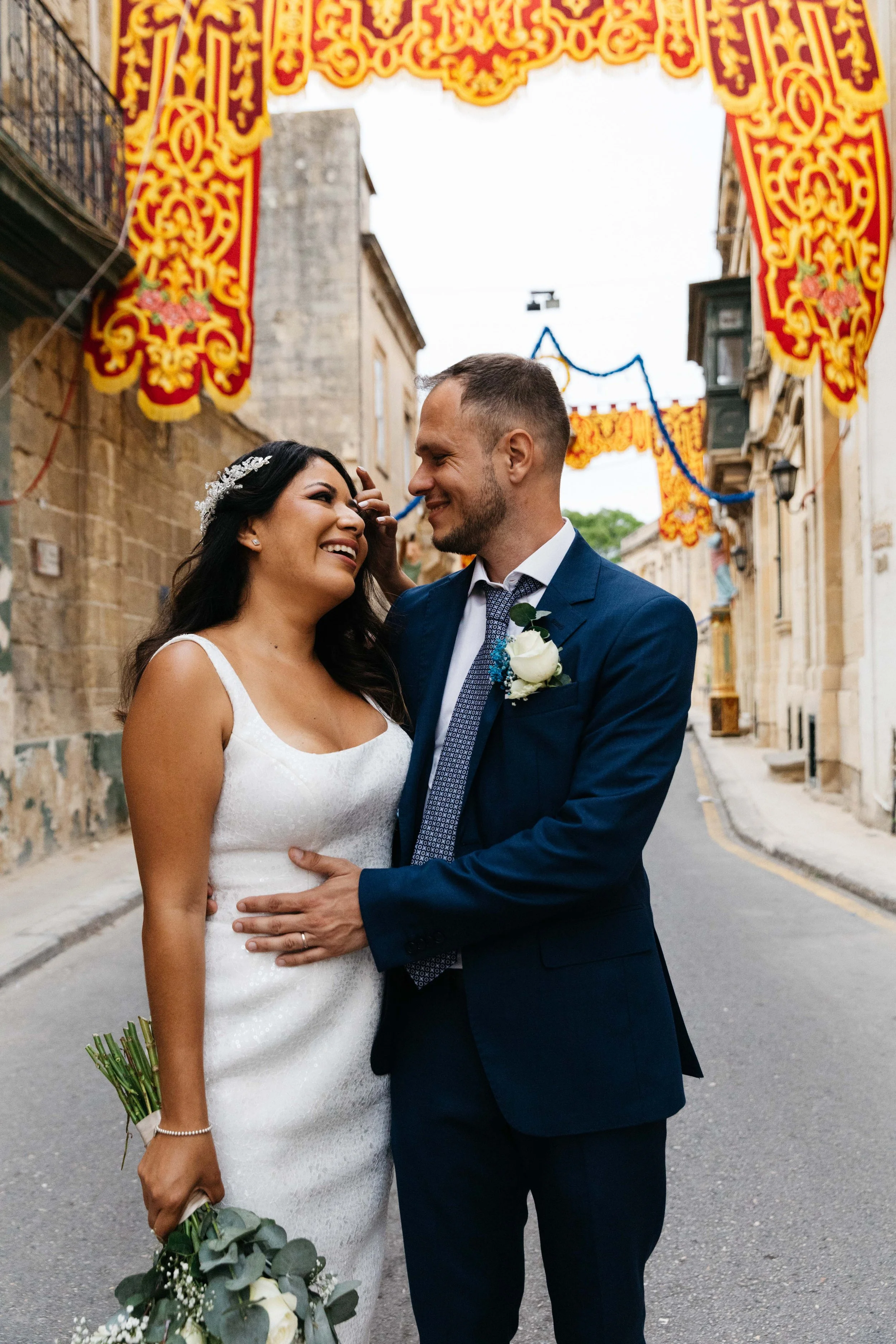 Bride and groom smiling on decorated street, holding bouquet.