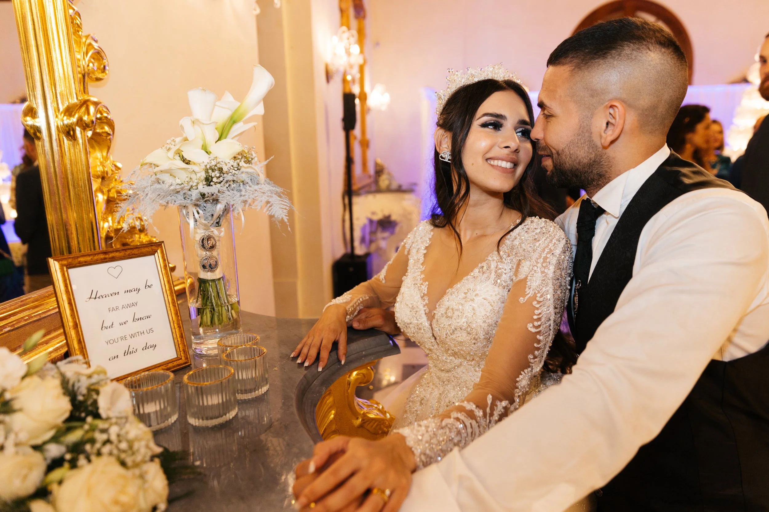 Bride and groom smiling at wedding reception, seated at a decorative table with flowers and a framed quote.