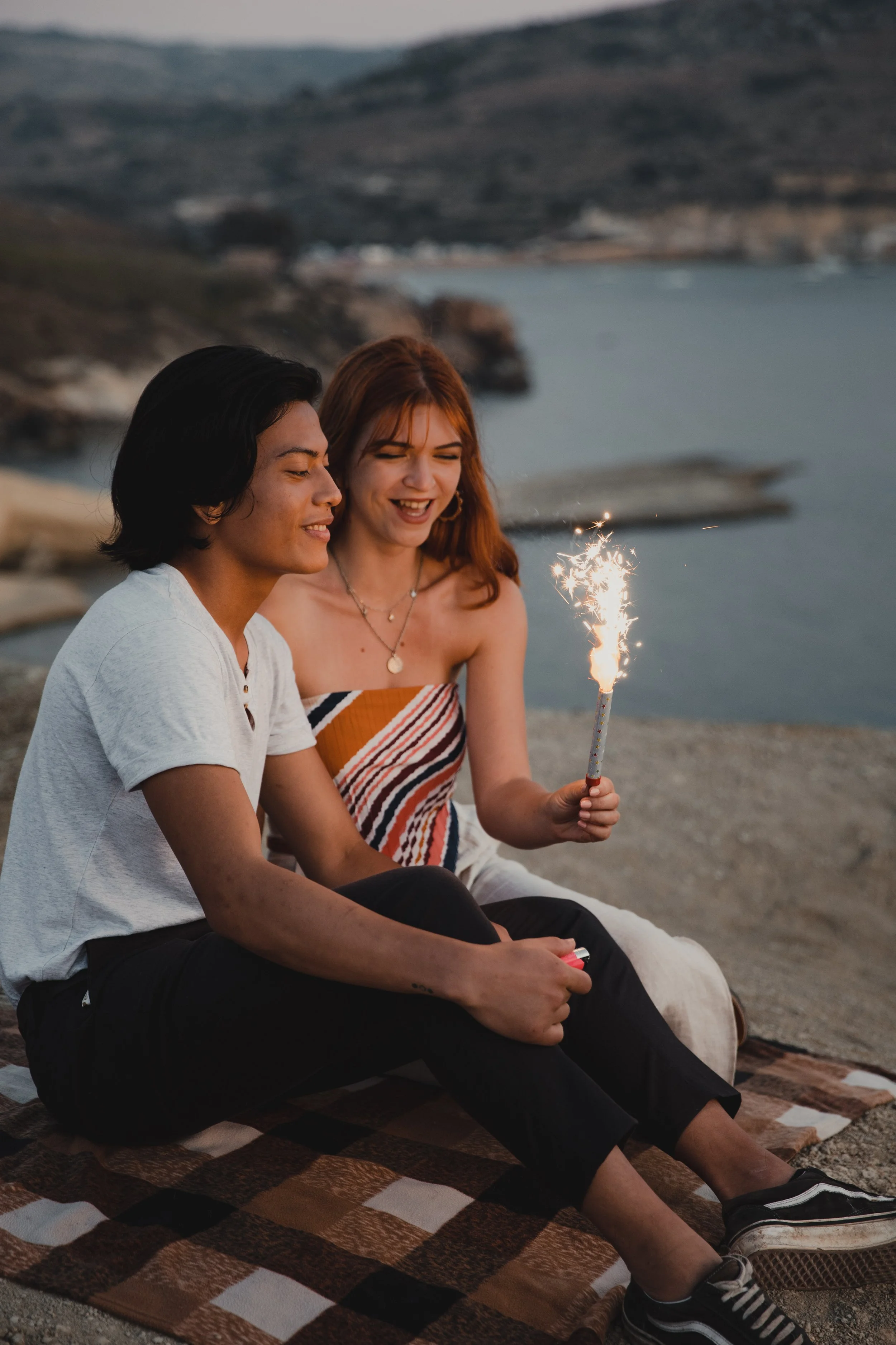 Two people sitting on a blanket by a lakeside, holding a lit sparkler, with hills in the background.