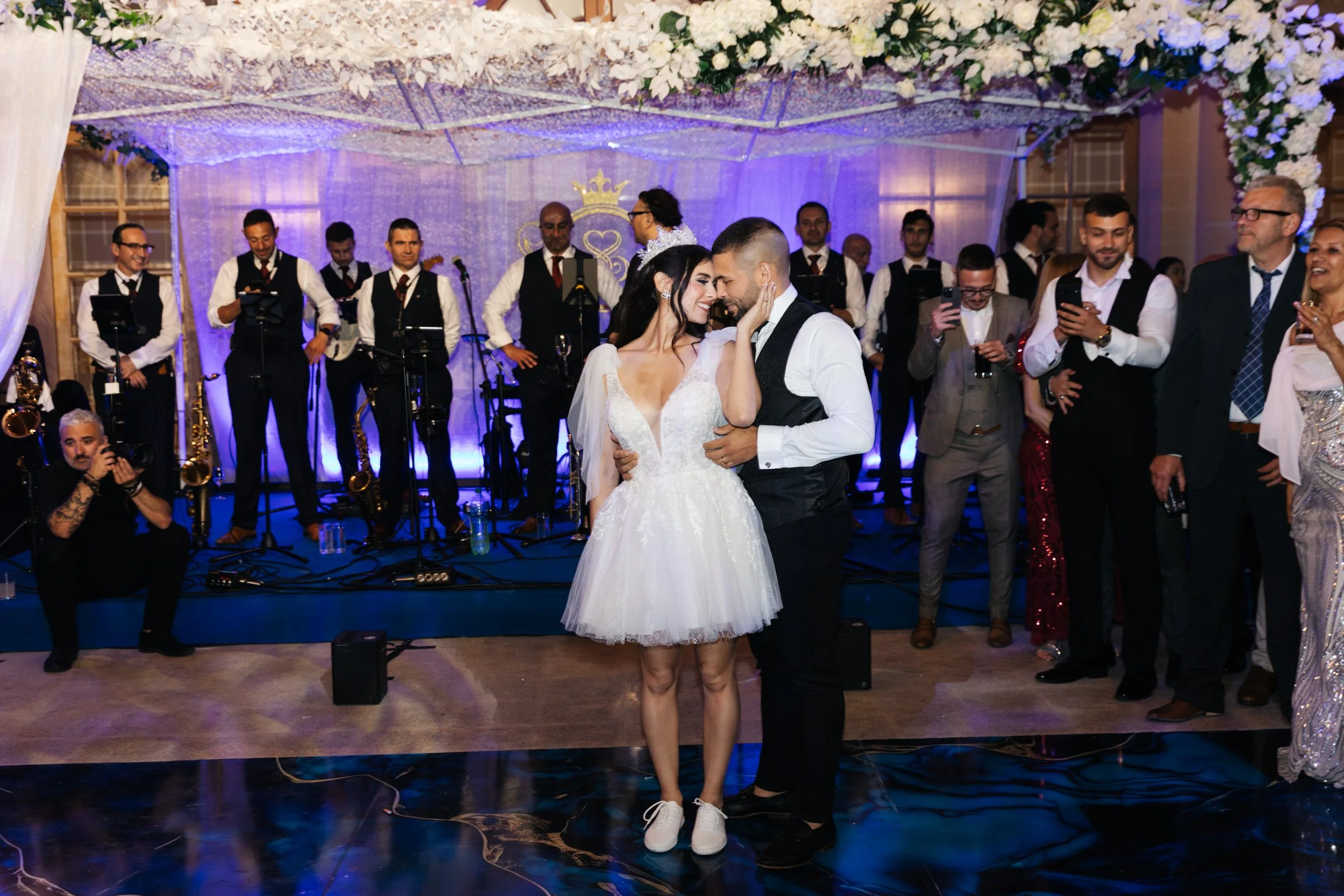 A wedding scene with a bride and groom dancing on a reflective floor, surrounded by guests. A live band with saxophones and brass instruments is performing in the background. There is white floral decoration above, creating a festive atmosphere.