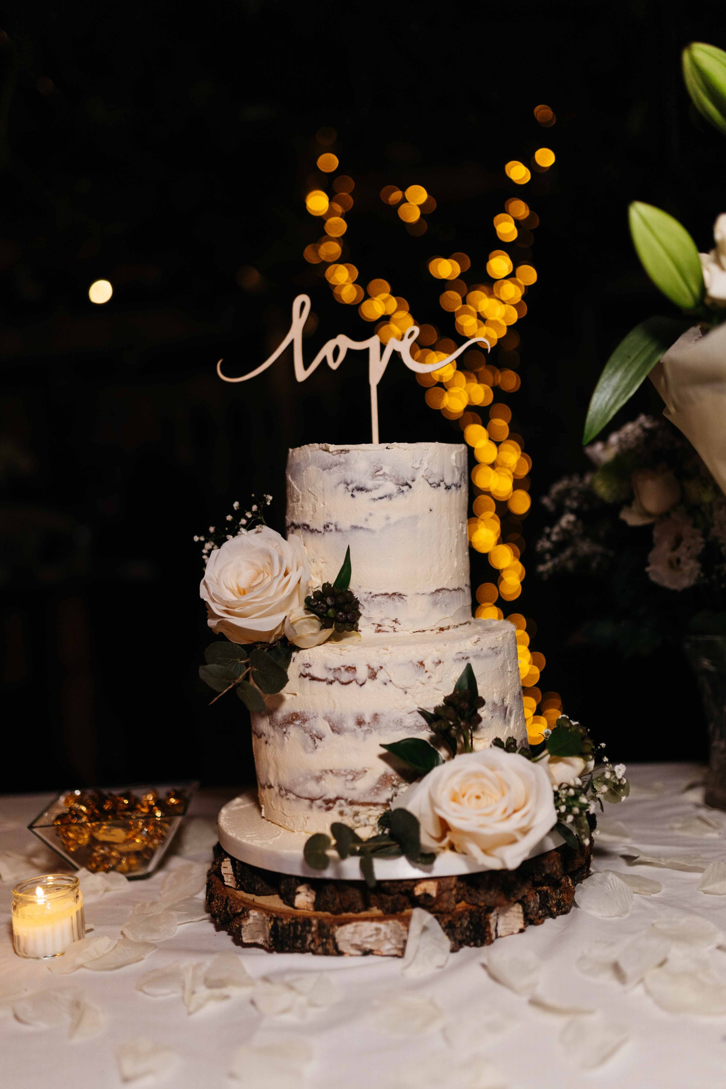 Rustic wedding cake with white icing, decorated with pale roses and greenery, topped with a 'love' sign, on a wooden stand with floral decorations and soft lighting.