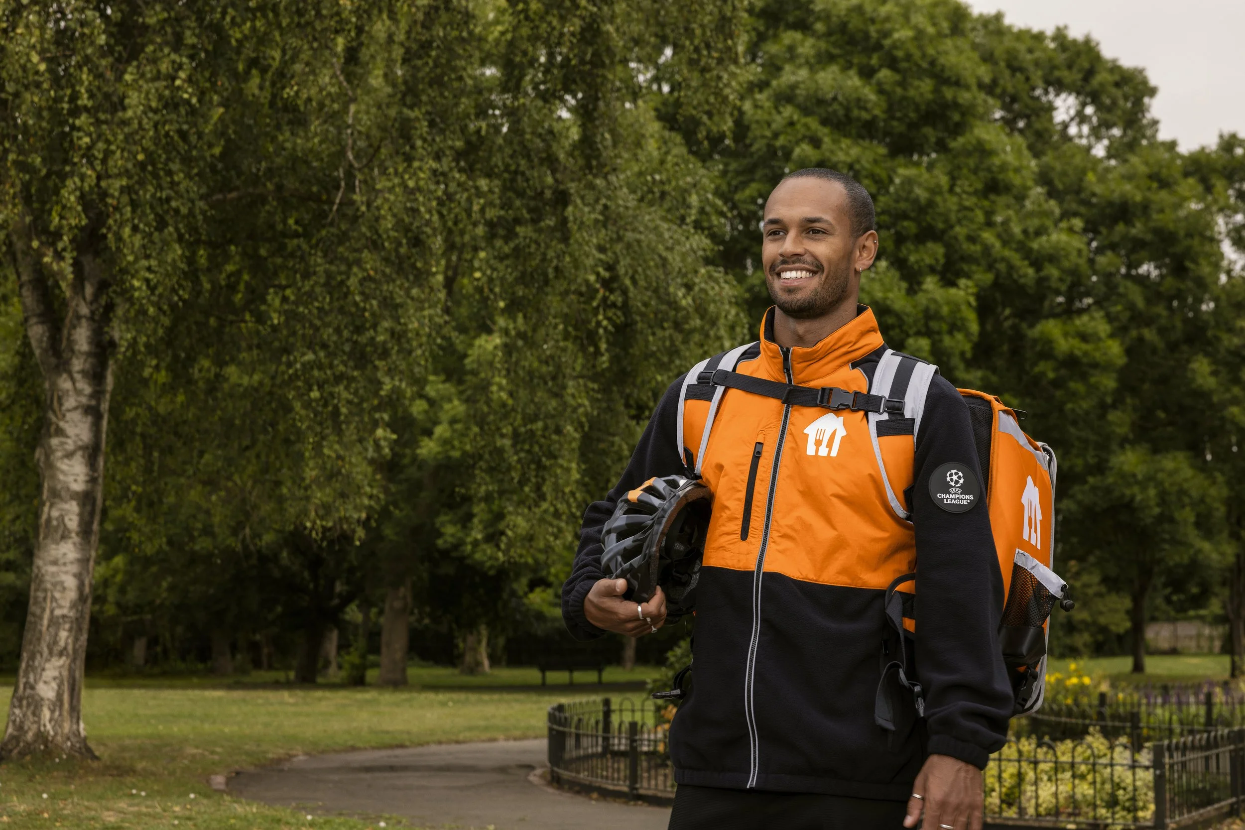 Just Eat campaign: a smiling courier with a helmet and branded delivery backpack in a park, photographed by Dean Bartolo.