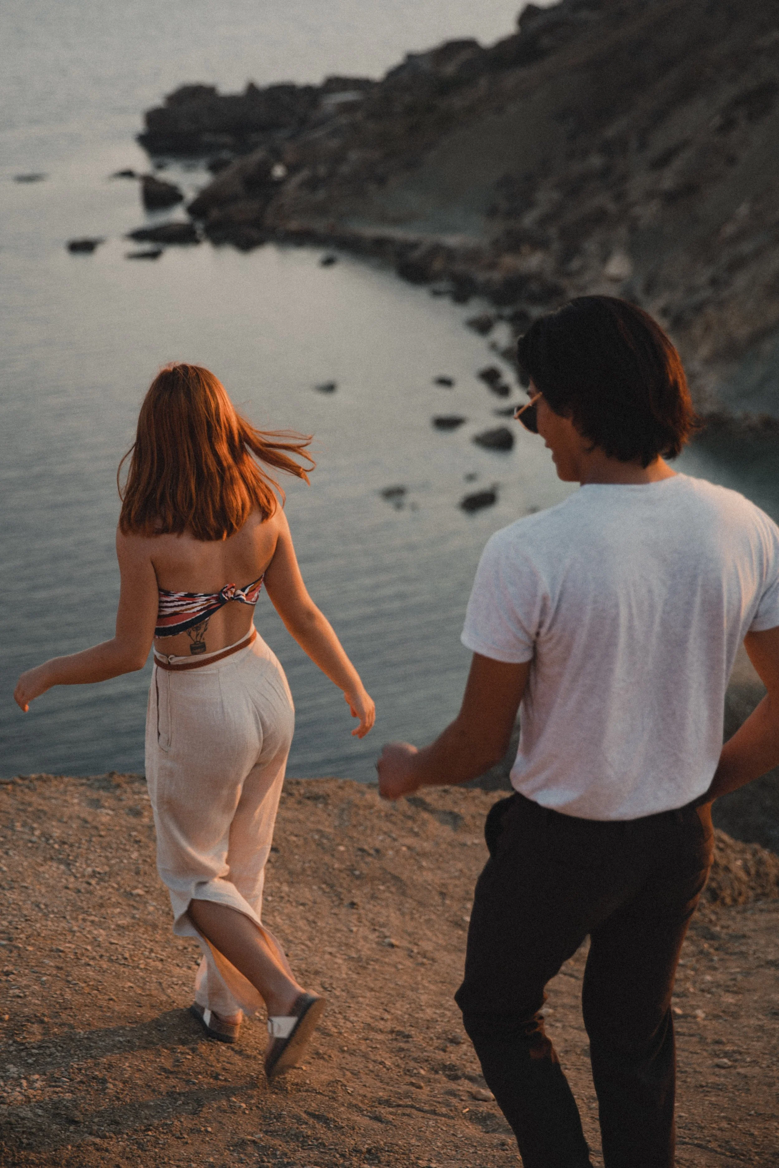 Two people walking on a cliffside overlooking the ocean at sunset.