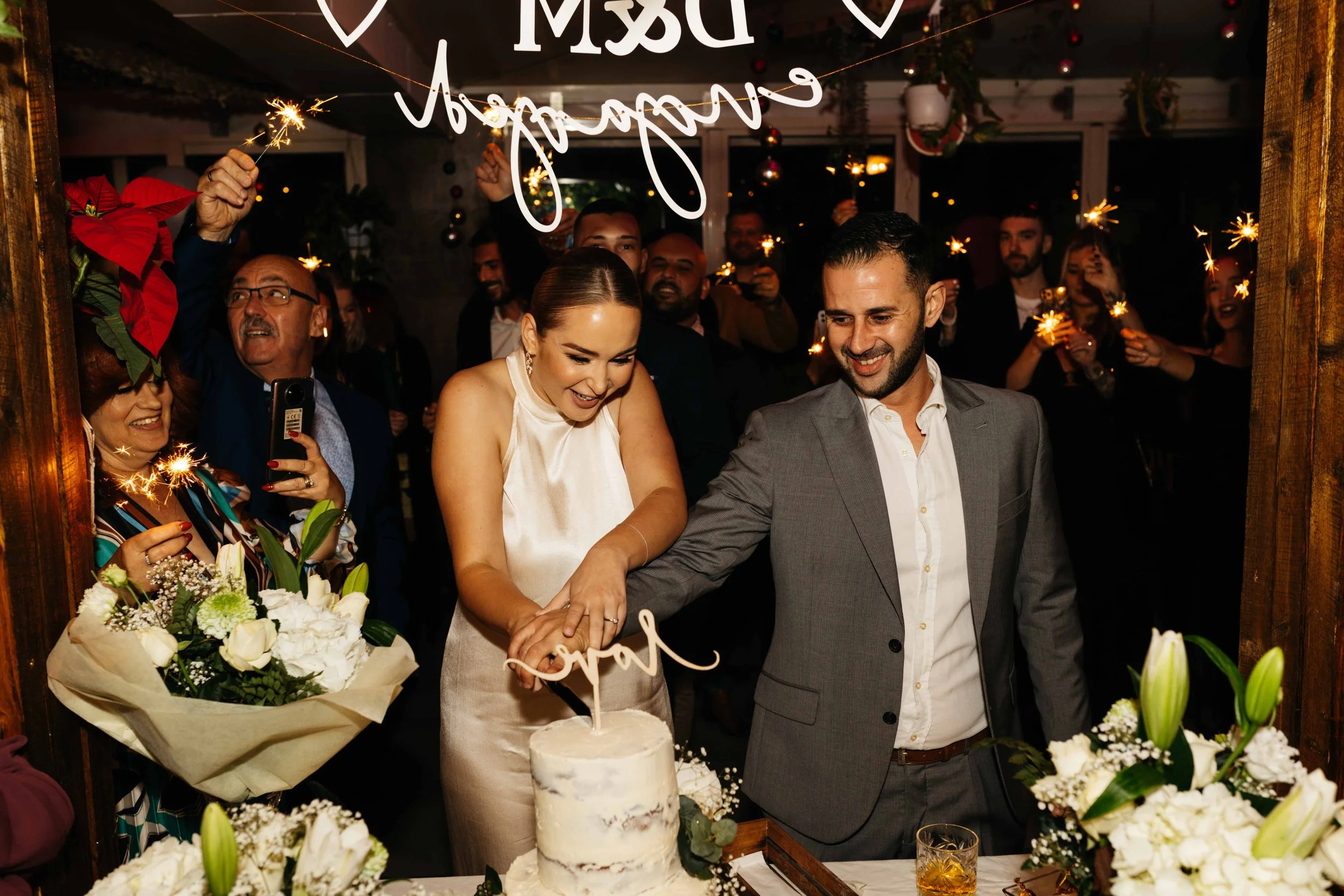 Couple cutting wedding cake surrounded by people holding sparklers at a celebration.