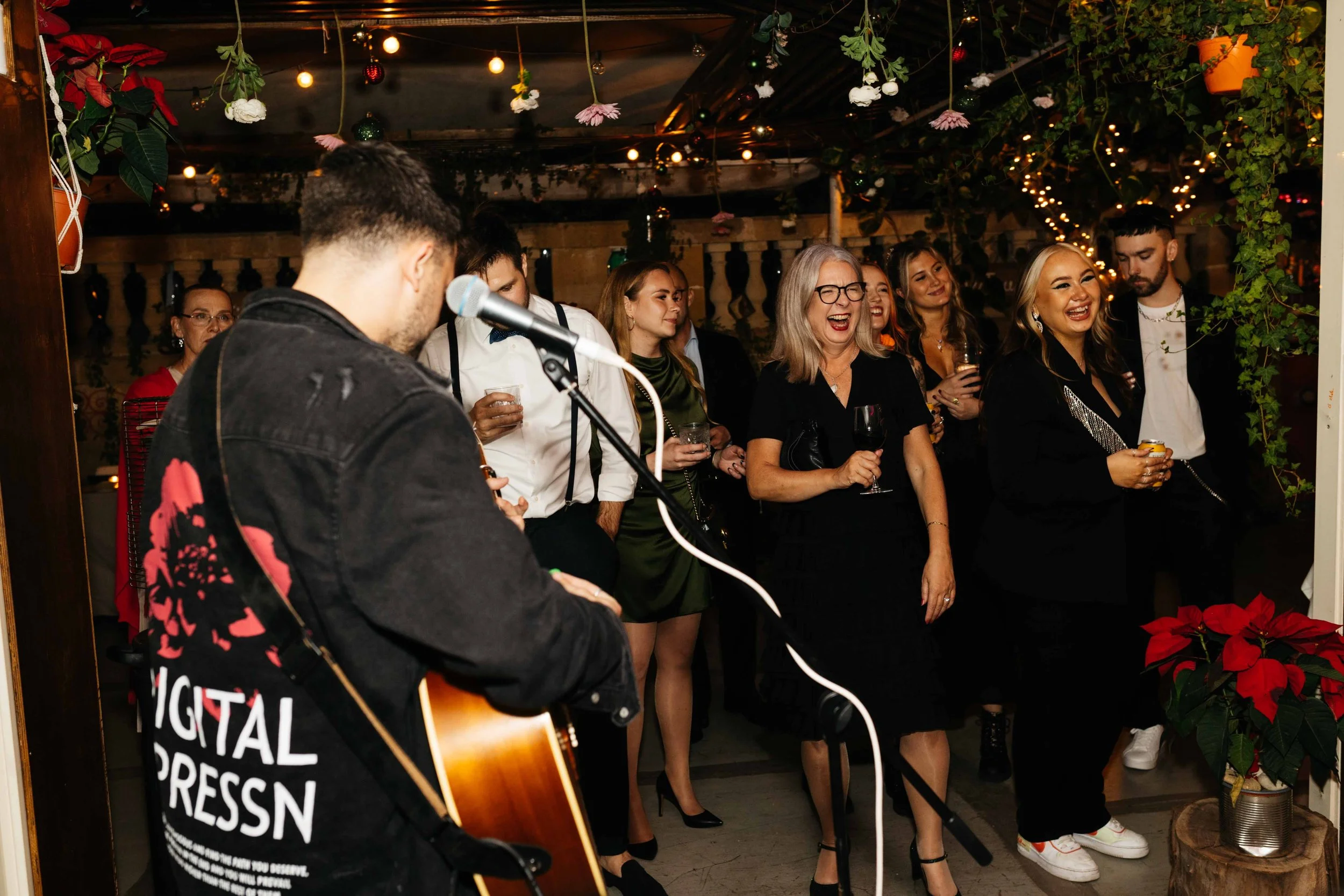 A musician performing in front of an audience at a festive indoor event, decorated with string lights and hanging plants. Attendees are holding drinks and smiling.