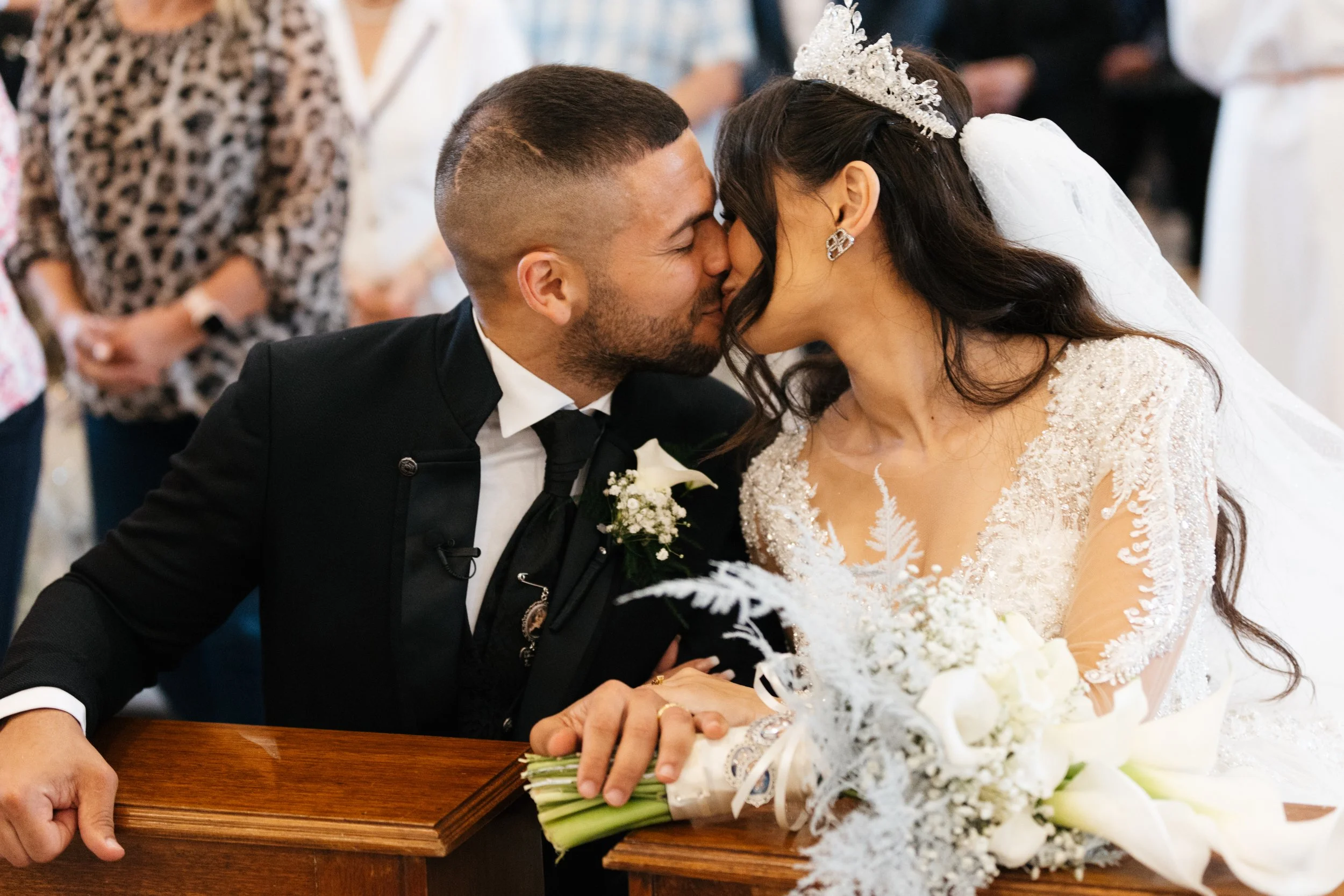 Bride and groom kissing at a wedding, the bride wearing a tiara and holding a bouquet, seated at a wooden altar.