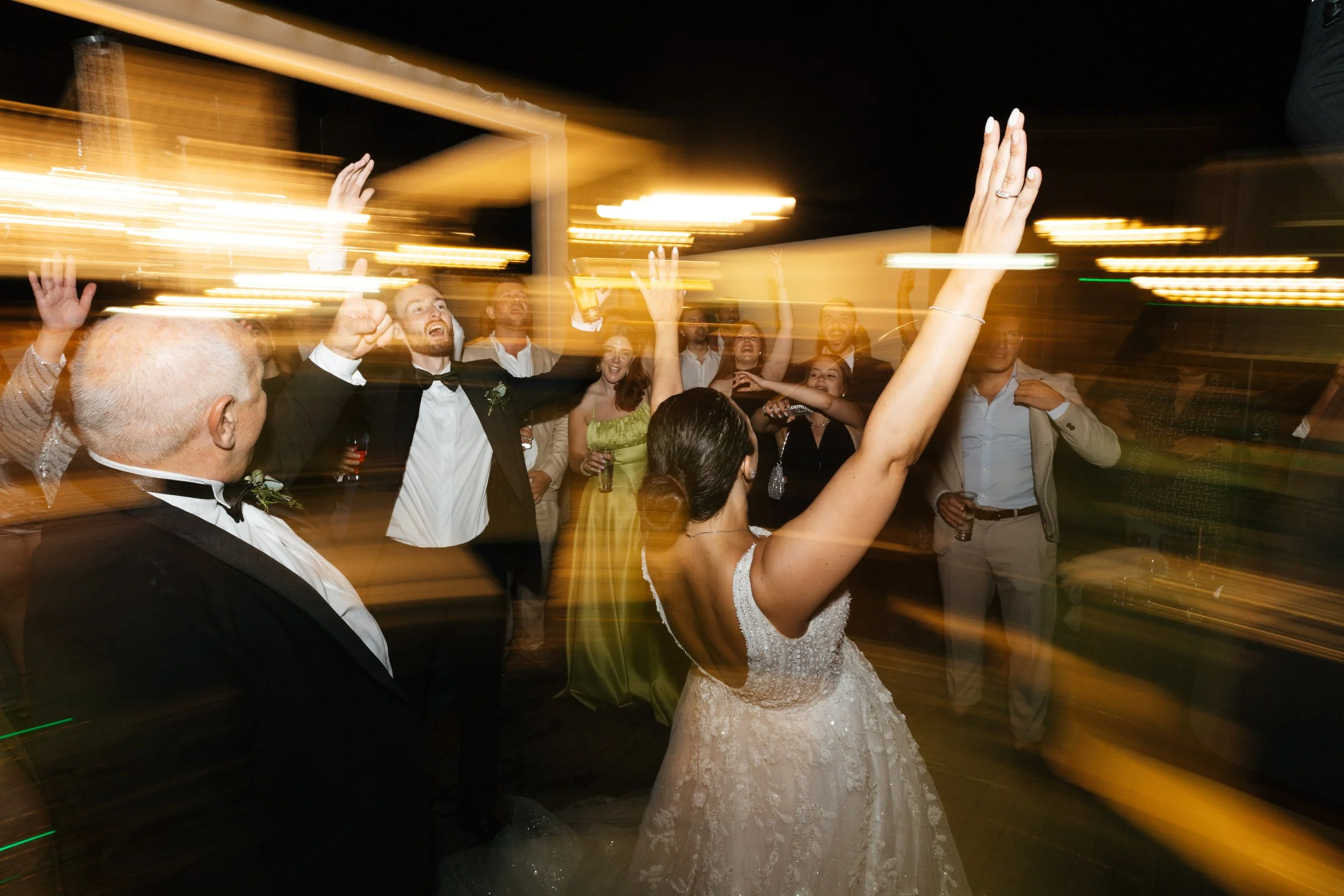 A group of people dancing and celebrating at a nighttime event, possibly a wedding reception, with colorful light streaks creating a dynamic effect. A bride in a white gown and a man in a tuxedo are visible in the foreground.