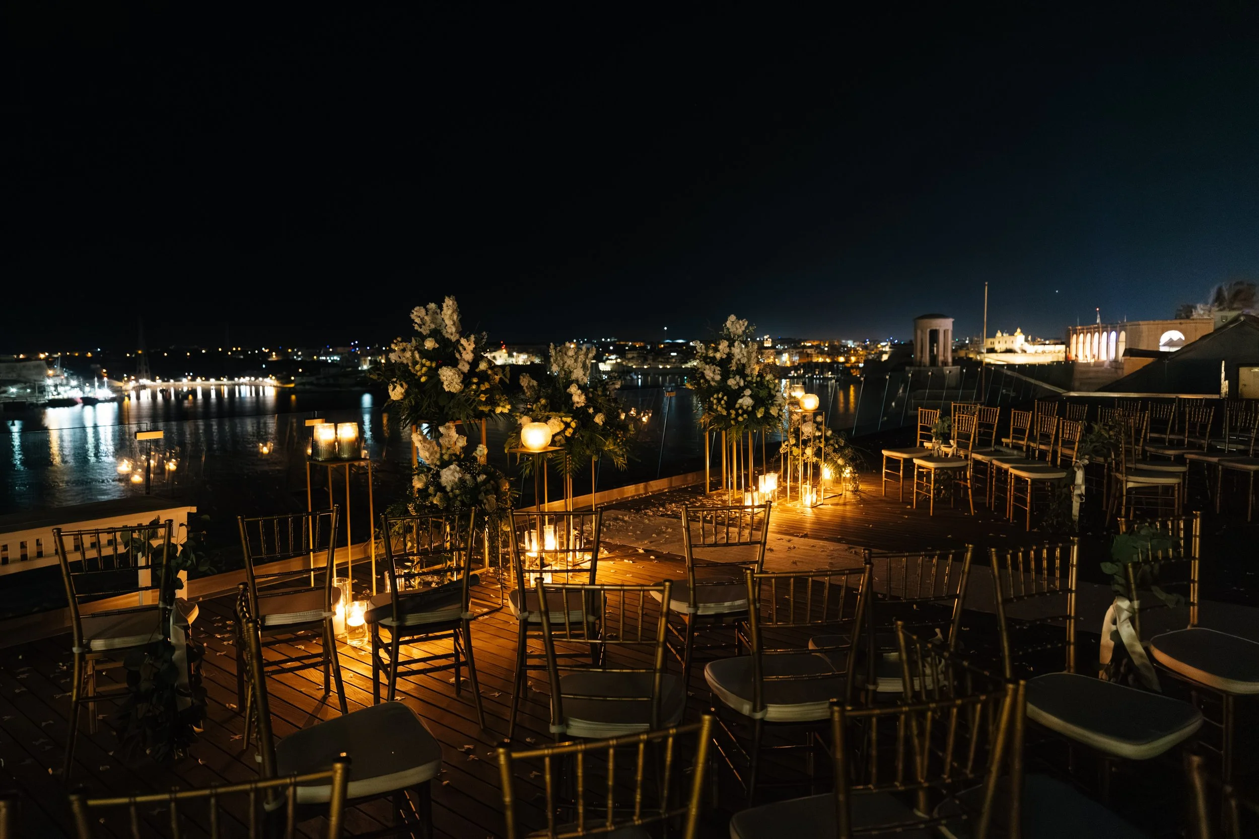 Nighttime outdoor wedding ceremony setup with chairs, floral arrangements, and lanterns overlooking a waterfront city skyline.