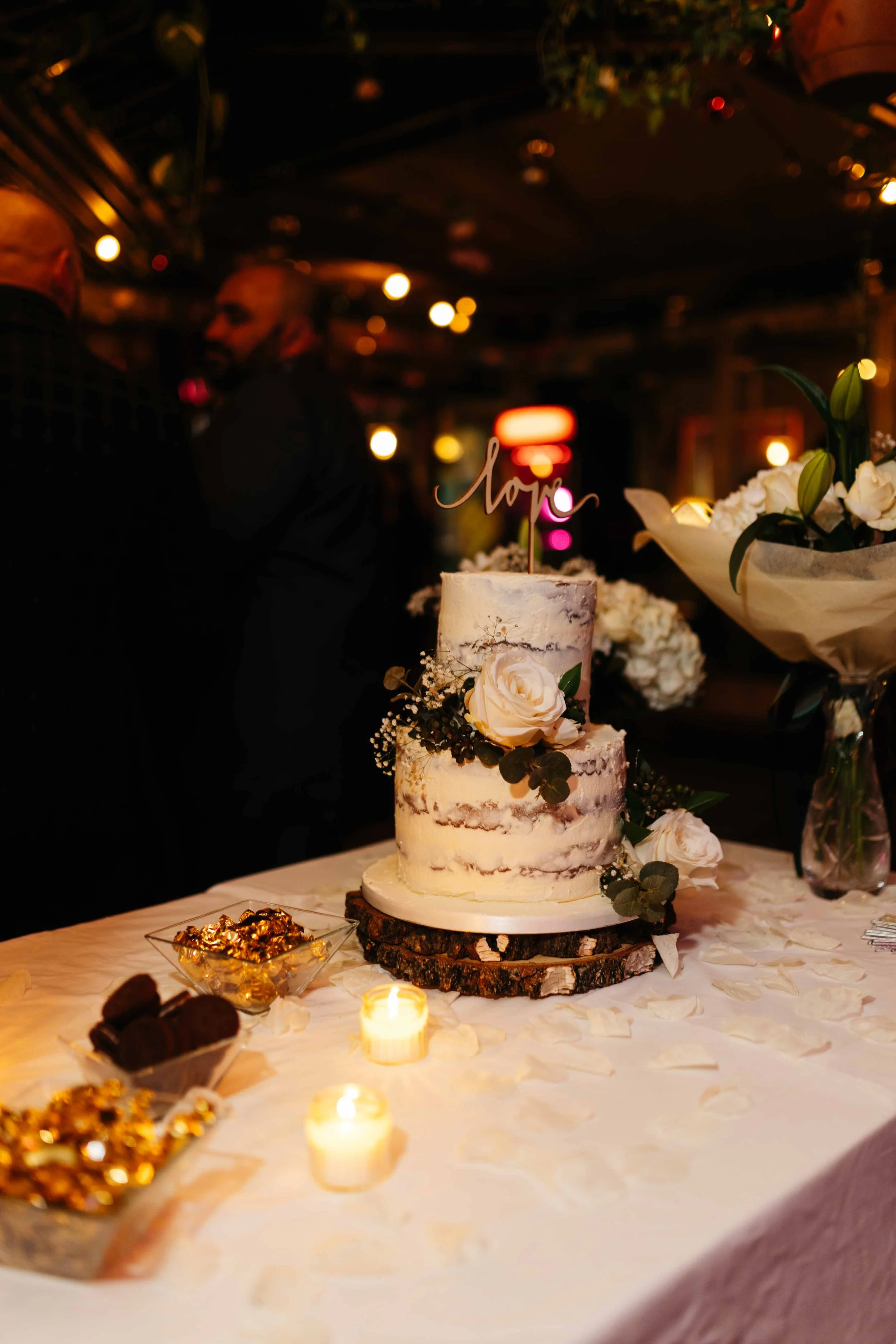 Three-tier semi-naked wedding cake with floral decorations and "love" topper, surrounded by candles and snacks on a decorated table.