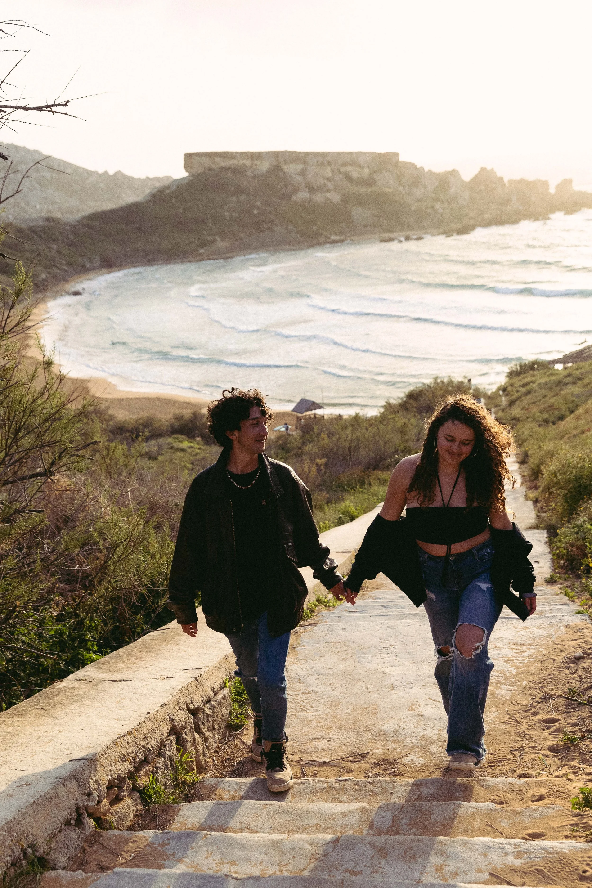 Couple holding hands walking up a path near a coastal beach.