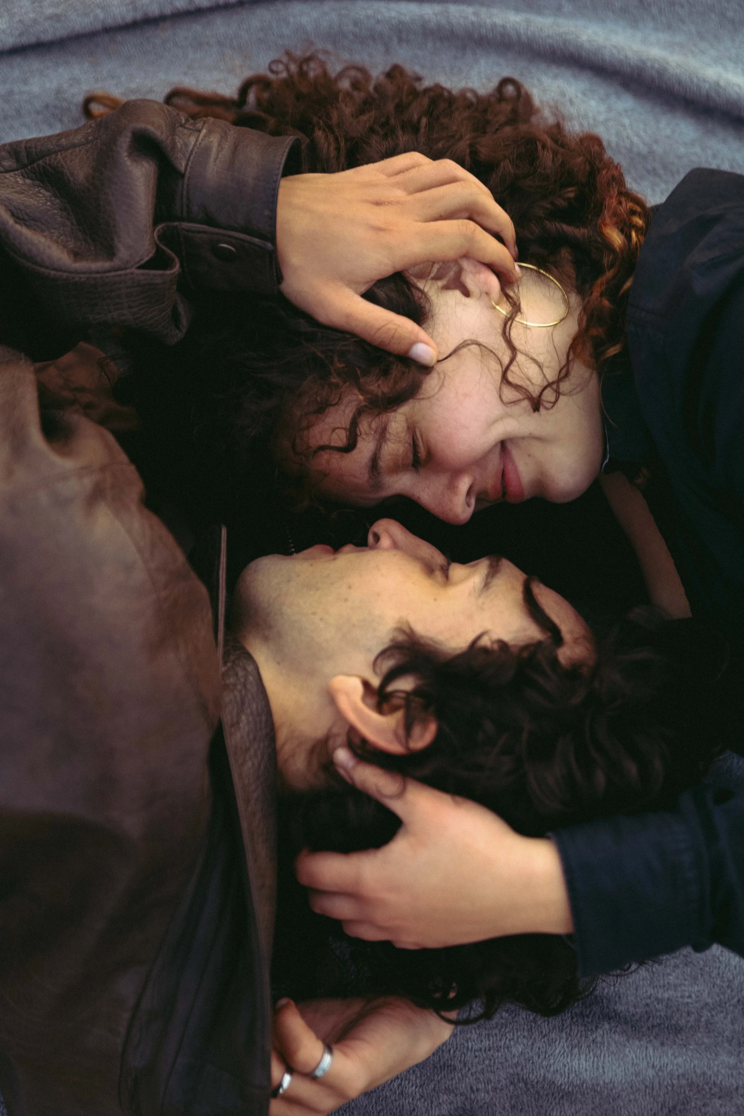 Couple lying close, faces touching, with curly hair and hands gently holding each other.