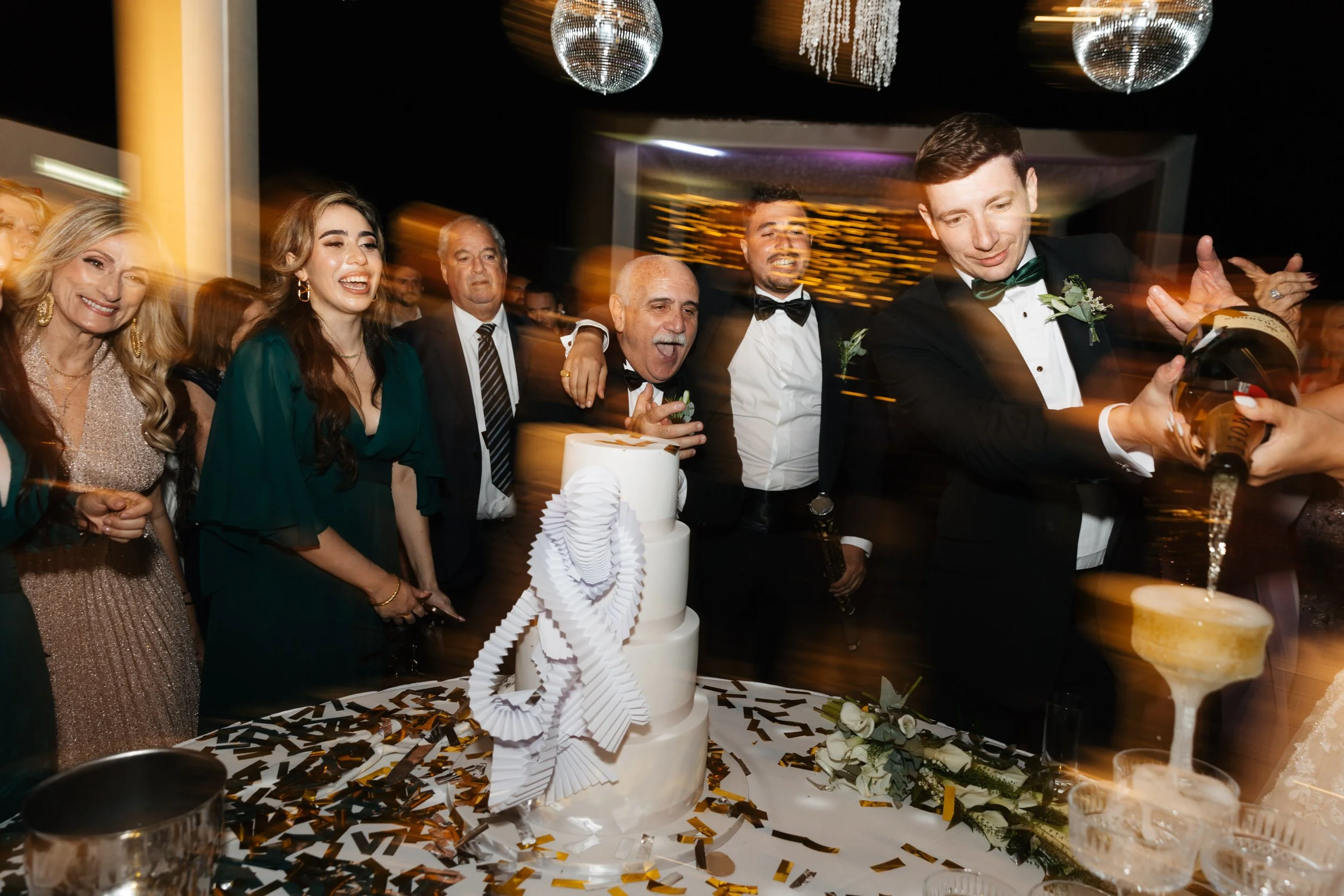 Group of people celebrating at an event, dressed in formal attire, with a white tiered cake on a table, and champagne being poured into a tower of glasses under disco balls.