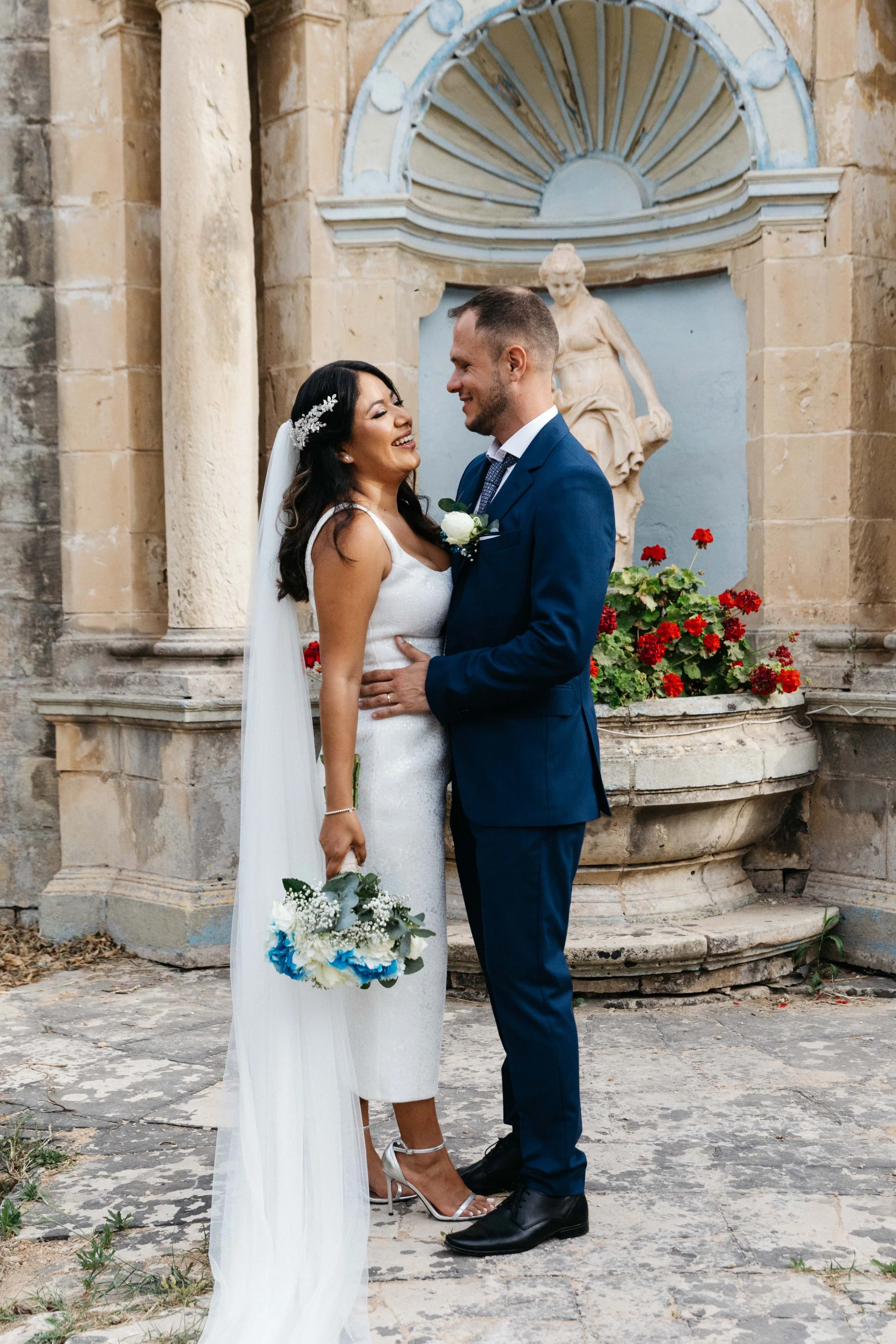 A bride and groom smiling at each other in front of stone architecture with a statue and flowers.