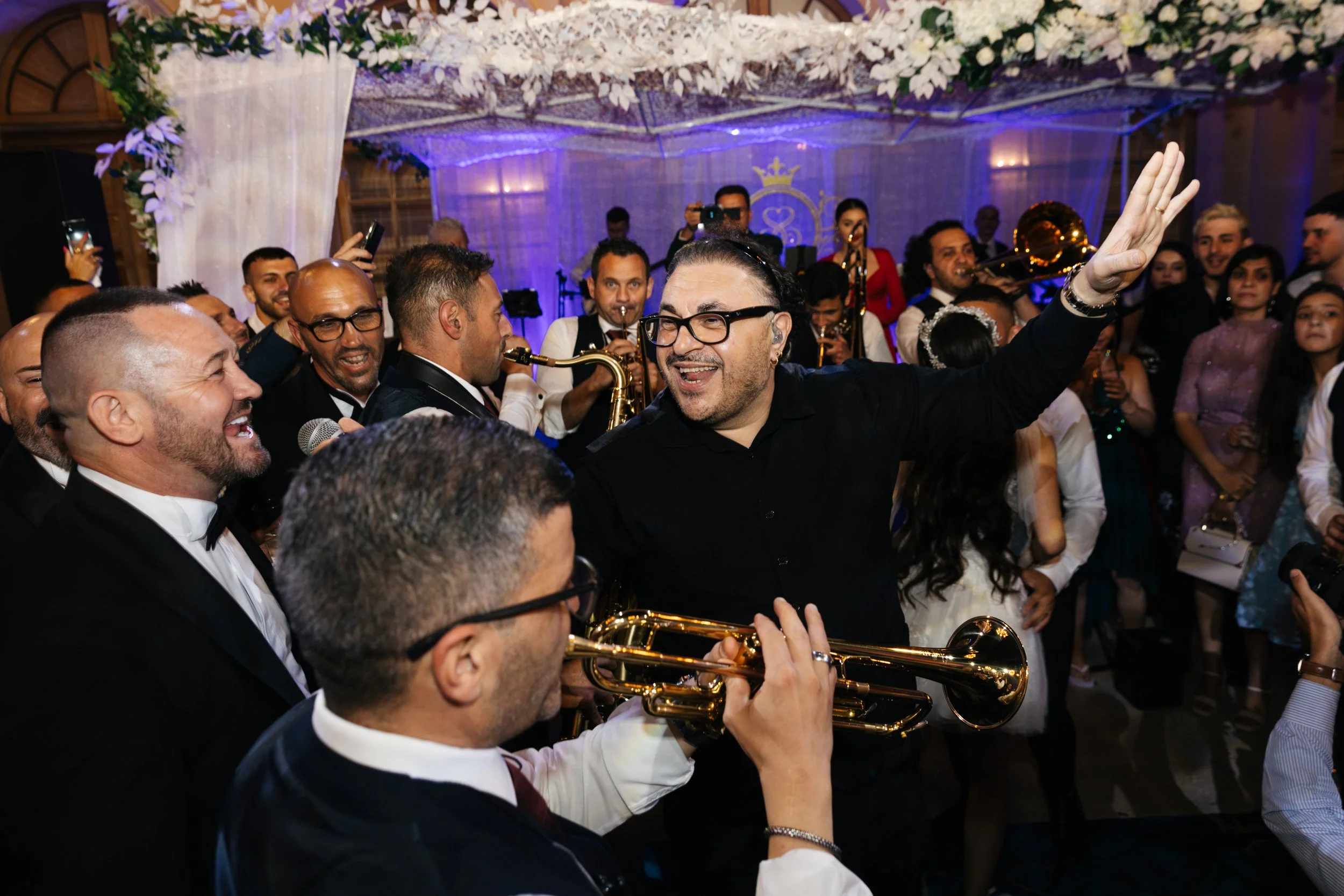 Group of people enjoying live music at a party with brass instruments, floral decorations in the background.