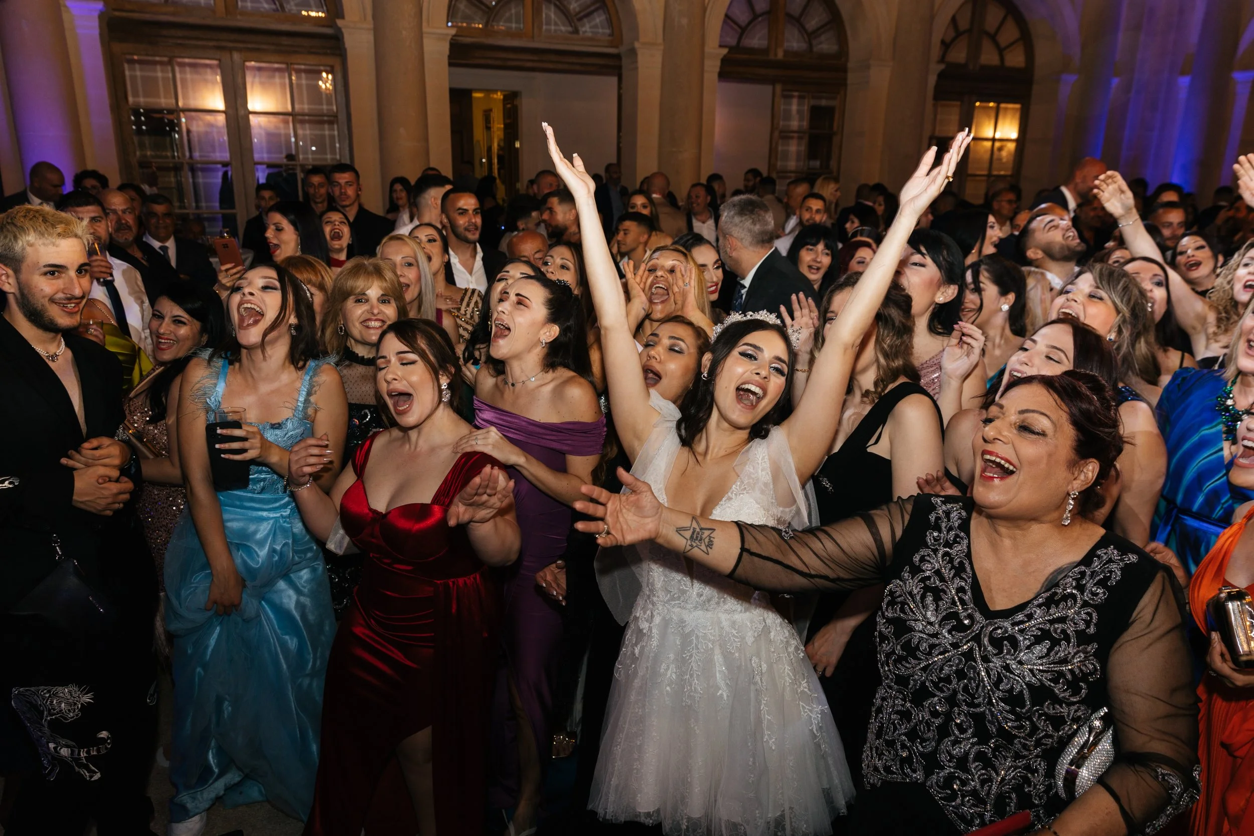 Group of people celebrating at a party, dressed in formal attire, including a bride in a white dress, with joyful expressions and raised hands.