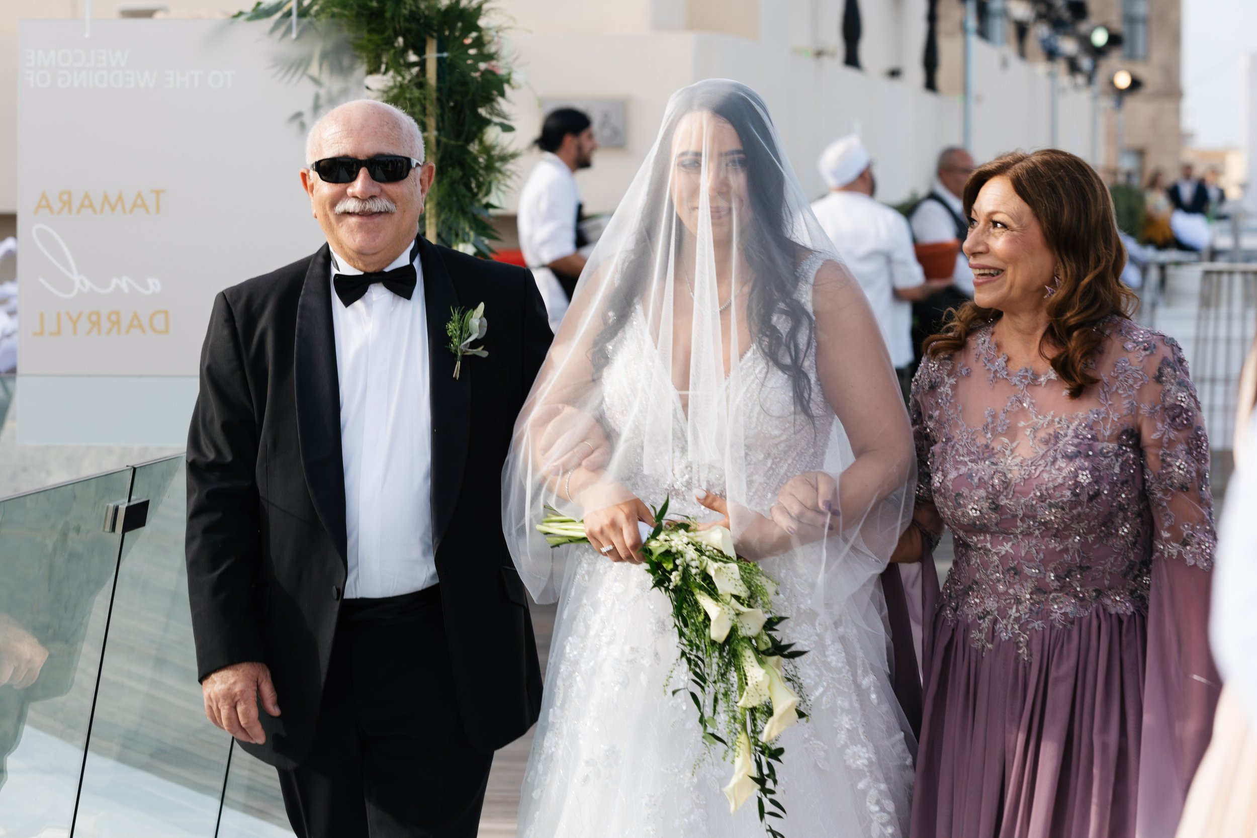 Bride in white gown with veil holding bouquet, flanked by a man in a tuxedo and a woman in a purple dress, at a wedding venue.