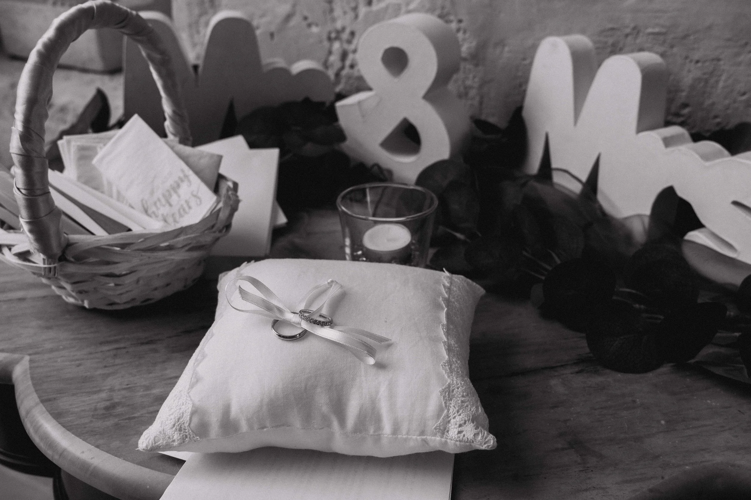 Black and white photo of a wedding setup with rings on a pillow, basket with cards, candle, and decorative 'Mr & Mrs' sign.