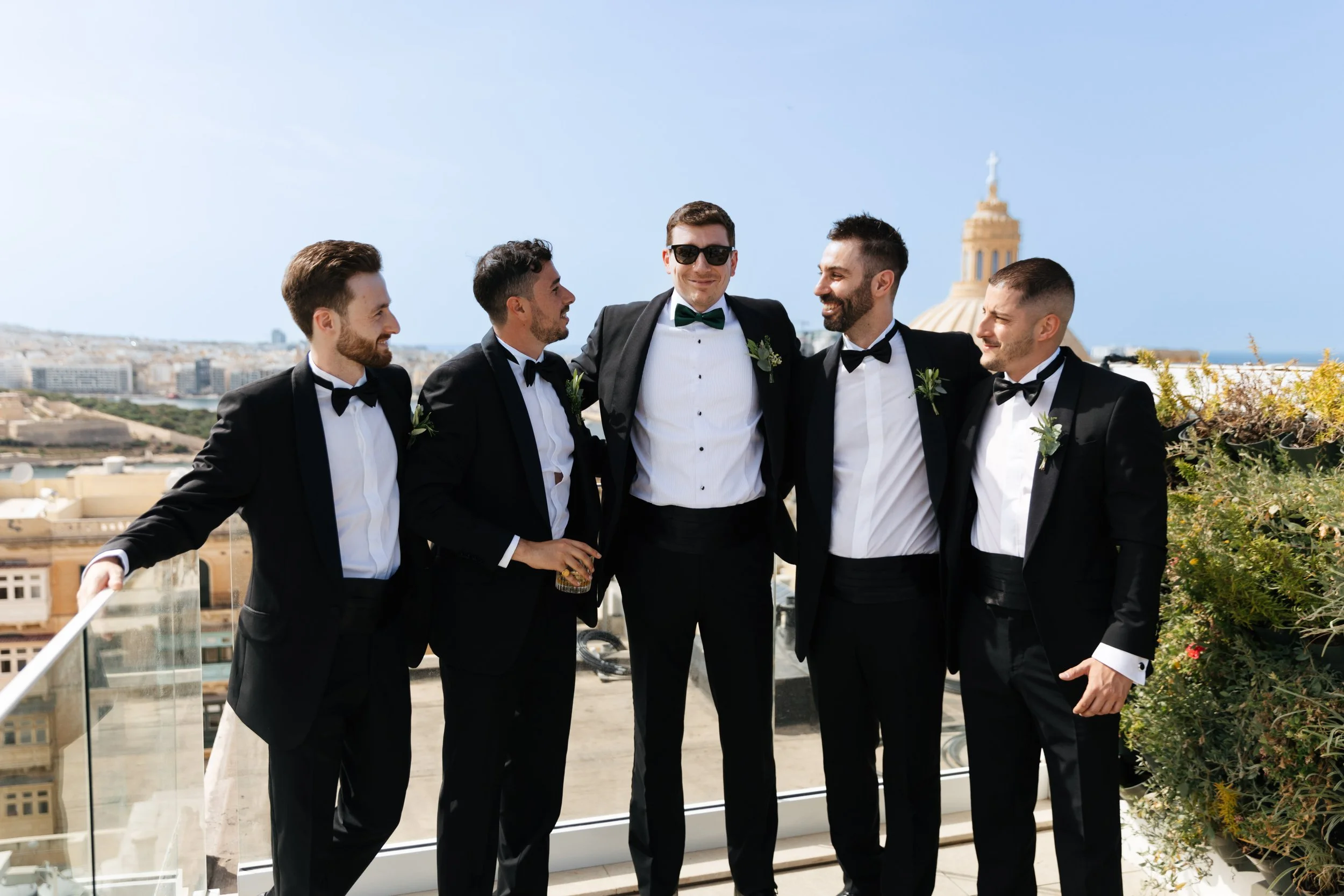 Five groomsmen in black tuxedos standing outdoors on a rooftop with a cityscape backdrop.