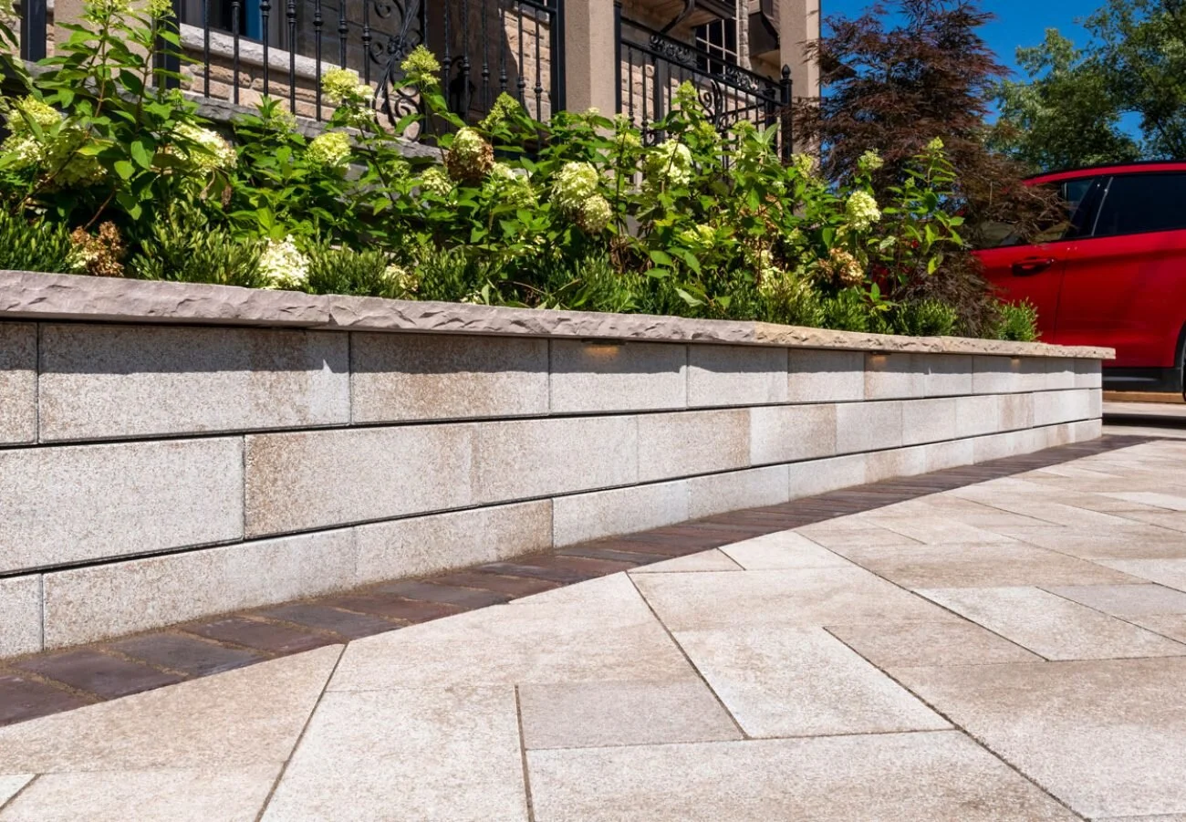 Decorative stone retaining wall with plants and flowers, paved pathway, red car partially visible, residential building in background.