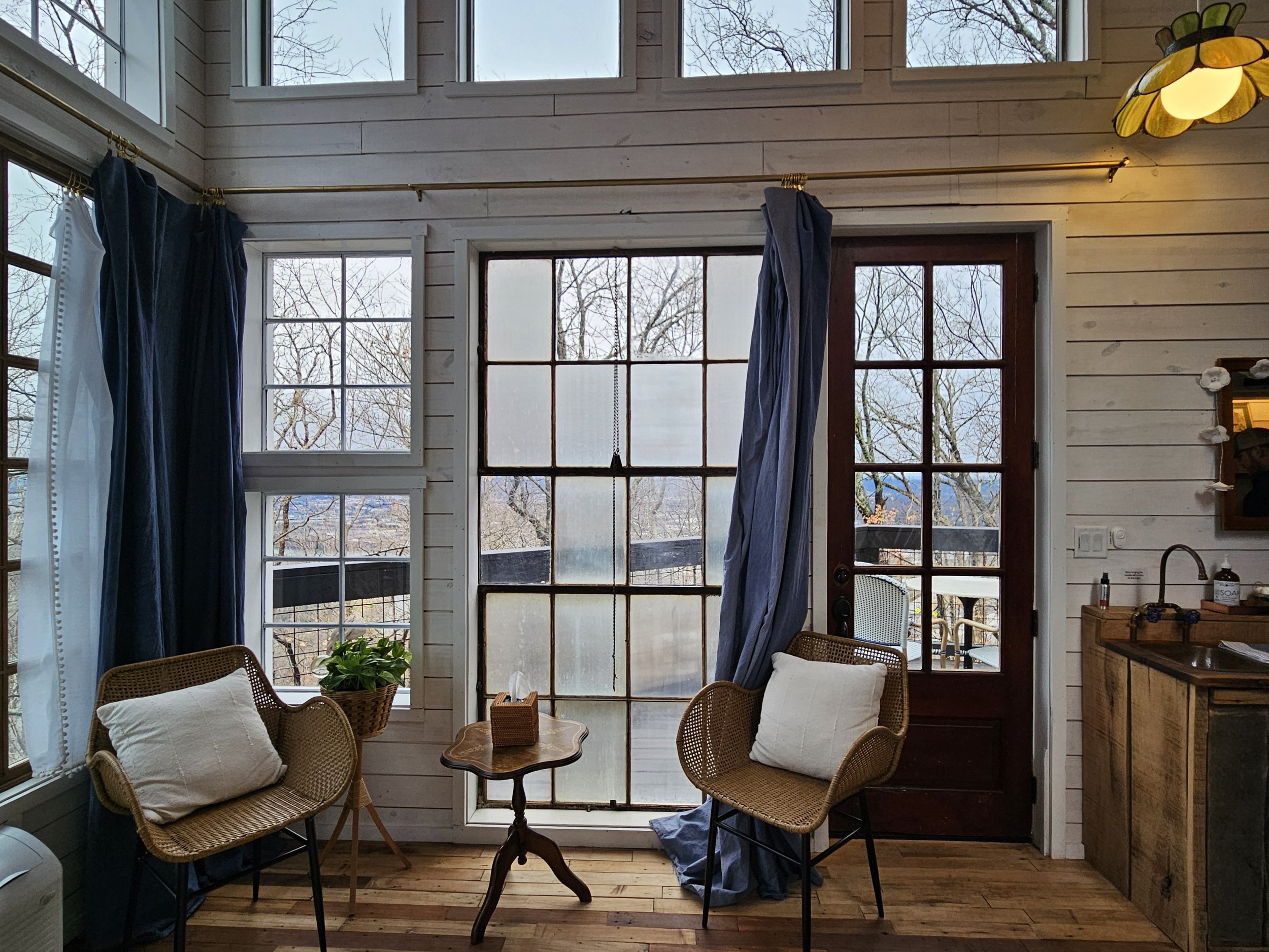 Interior of a cozy room with large windows, two wicker chairs with cushions, a small wooden table with tissue box, potted plant, and a sliding glass door leading outdoors. There's a wooden cabinet with a sink on the right and a stained glass ceiling light. Trees are visible outside.