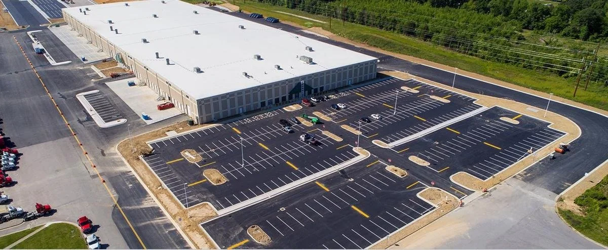 Aerial view of a large sortation center with a newly paved parking lot with marked spaces, surrounded by greenery and additional parking areas.