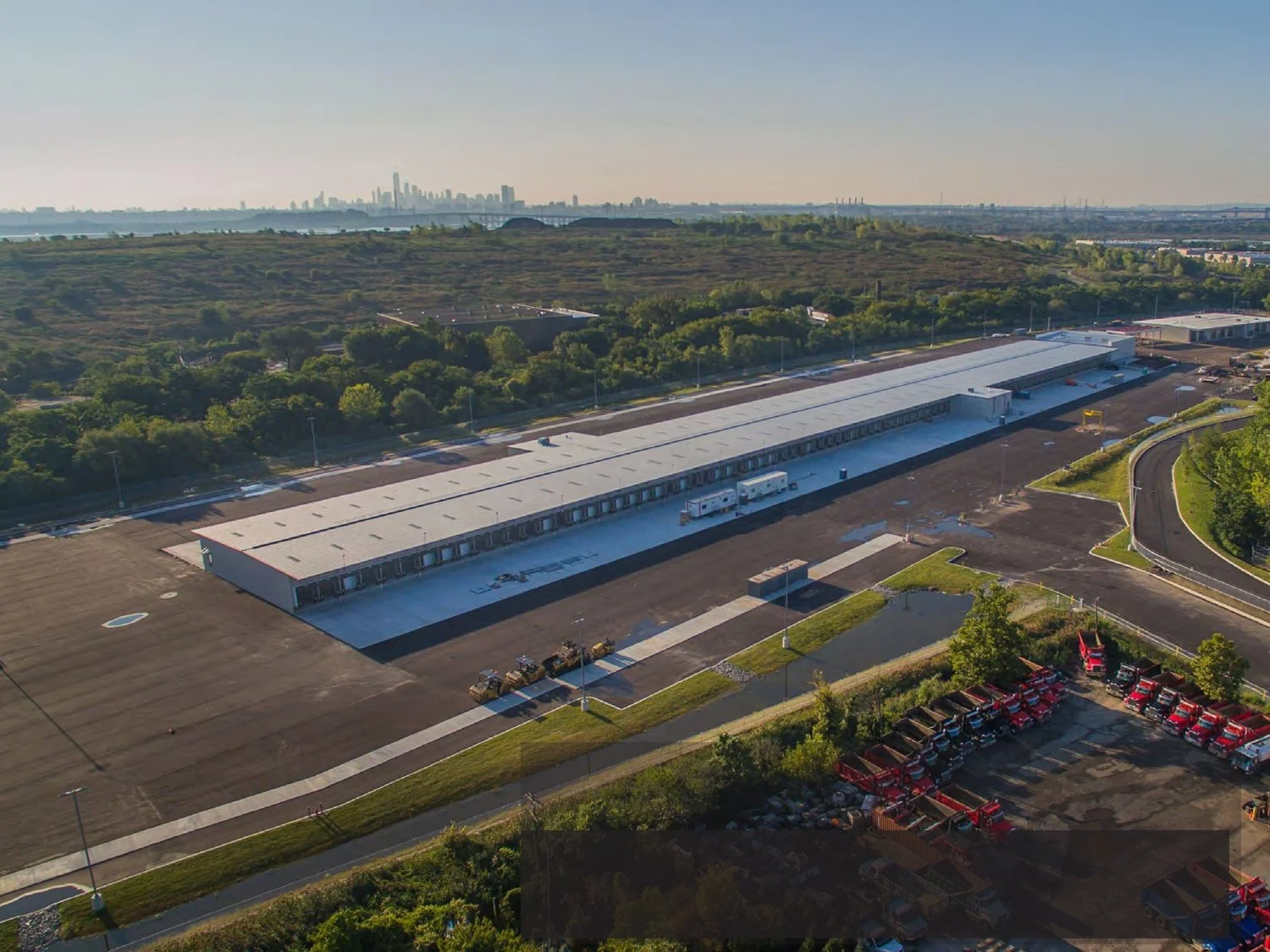Aerial view of a large industrial truck terminal with multiple loading bays, a parking lot with red trucks parked, and surrounding greenery under a clear sky, with city skyline in the distance.