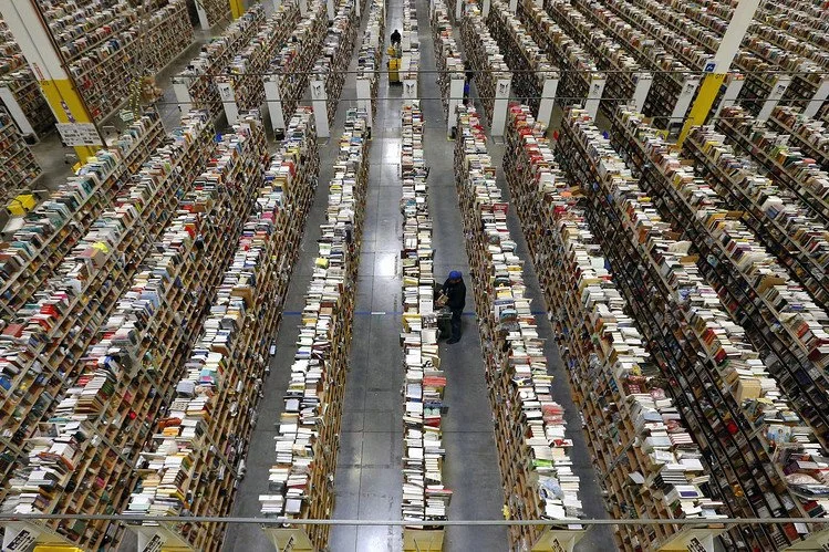 A person walking inside a large sortation center with tall bookshelves filled with books on multiple aisles.