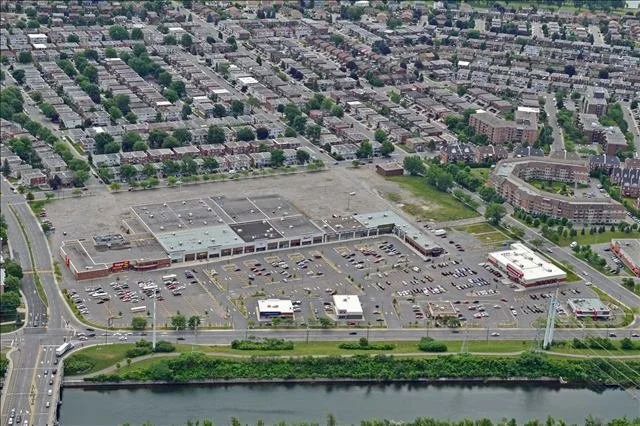Aerial view of a large shopping mall with parking lots, surrounded by residential neighborhoods, near a river and a highway.