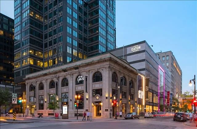 Street view with historic stone building and modern skyscrapers near Eaton Center in an urban area of Montreal at dusk, with cars and pedestrians.