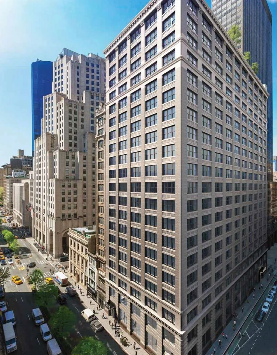 Tall Manhattan city building with many windows and a beige facade, surrounded by other skyscrapers and a busy city street below with cars and pedestrians.
