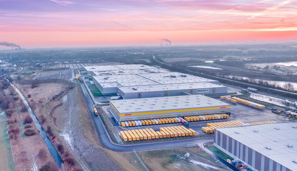 Aerial view of Amazon and DHL logistics warehouses with trucks parked outside during sunset.