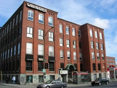 Brick building with 'Ubisfot' signage at the top, multiple windows, Saint-Laurent street in Montreal with cars, and pedestrians.
