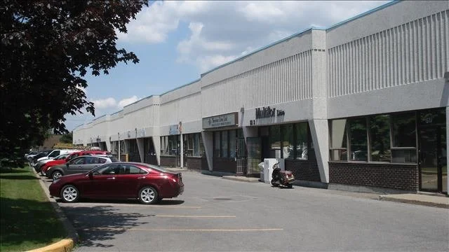 Shallow Bay shopping plaza with parked cars in front, storefronts with signs, and a partly cloudy sky.