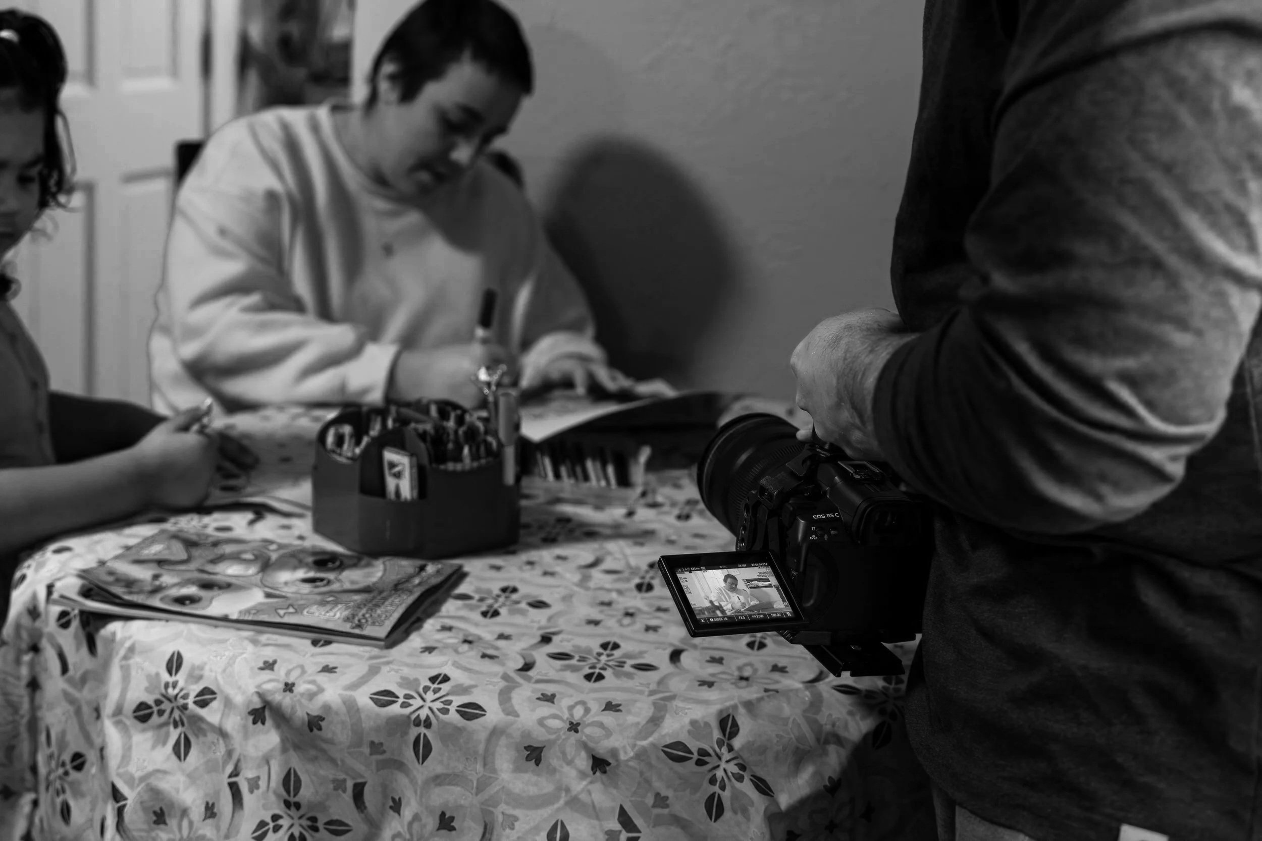 Close-up of a person filming children at a table, with children engaged in activities and magazines, in a black and white photo.