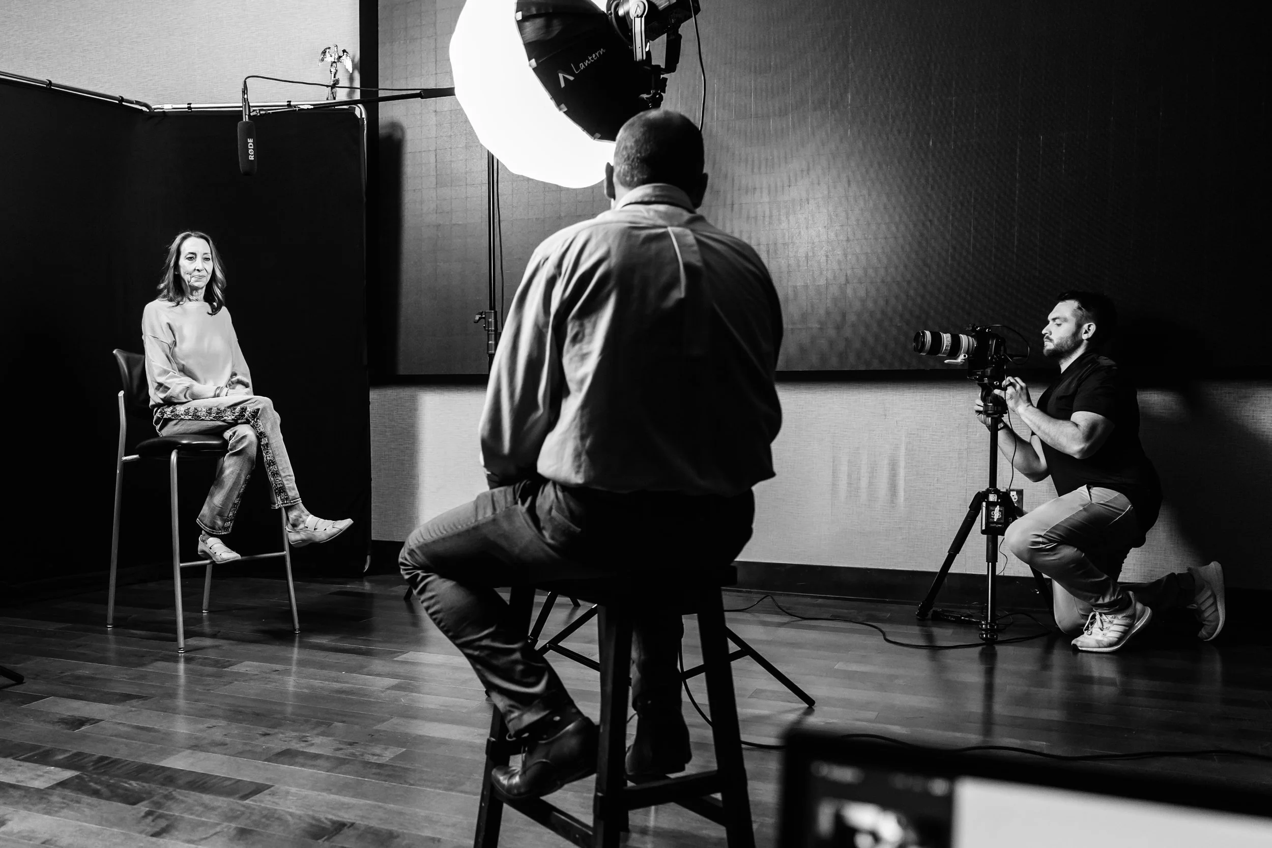 A woman seated on a high chair in a recording studio or interview setting, with a microphone hanging above her, being filmed by a cameraman kneeling with a camera, and another person sitting on a stool observing or directing, with professional lighting illuminating the scene.
