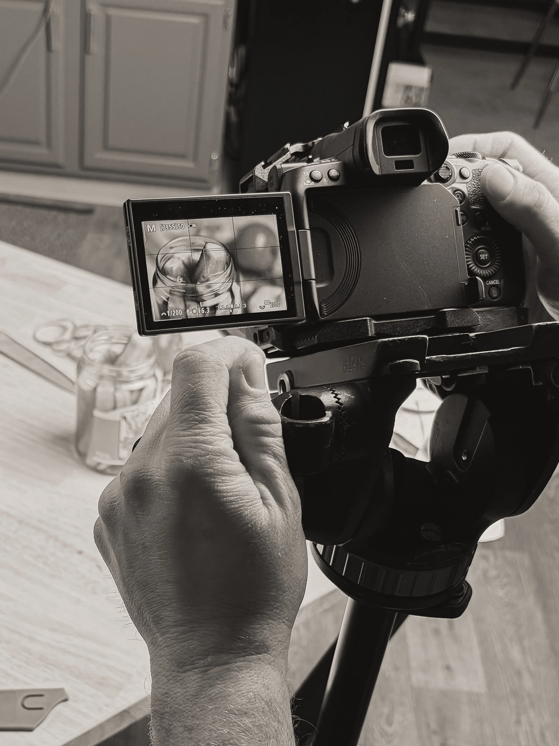 A person holding a camera on a tripod, taking a photo of a jar of pickles on a wooden table.