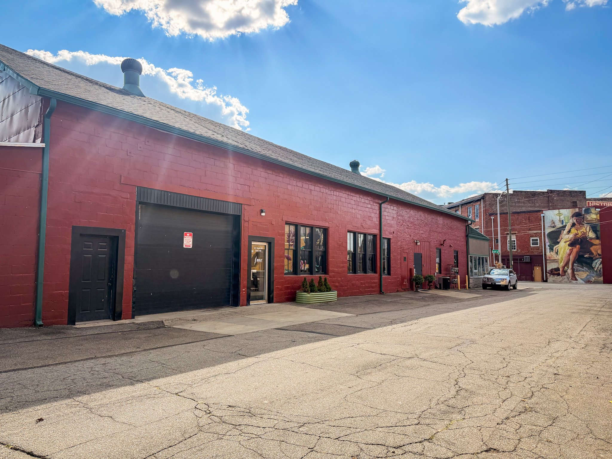 A red brick building with a black garage door, a door with a window, and several windows, with solar panels on the roof. The building is in an alley with a cracked pavement. A car is parked near the building, and mural art can be seen on the wall in the background.