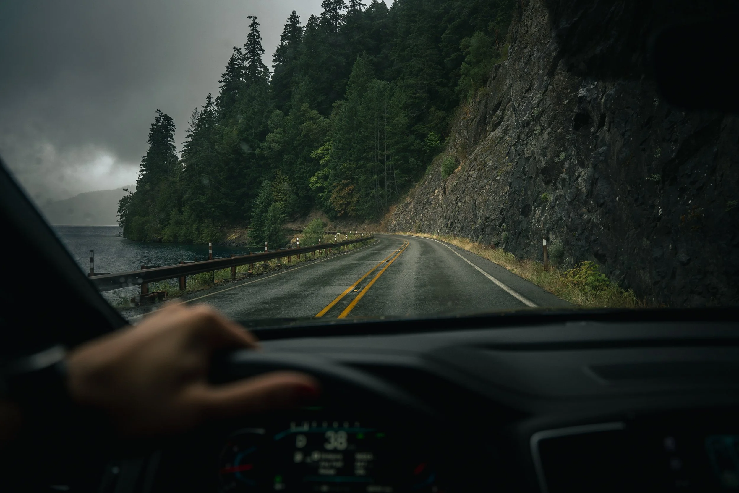 A car is driving along a winding road next to a body of water, with a steep, rocky hillside covered with dense green pine trees on the right. The weather appears to be overcast and misty.