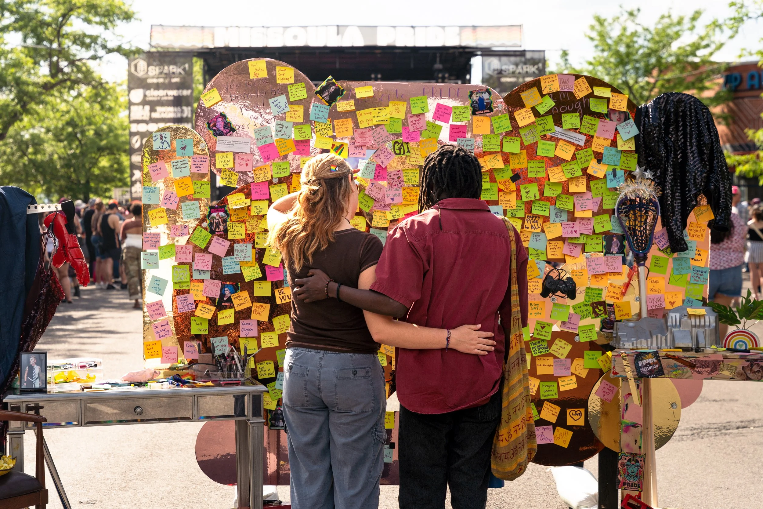 A couple holding each other in front of a large display covered with colorful sticky notes at an outdoor event with many people in the background, trees, and a stage.