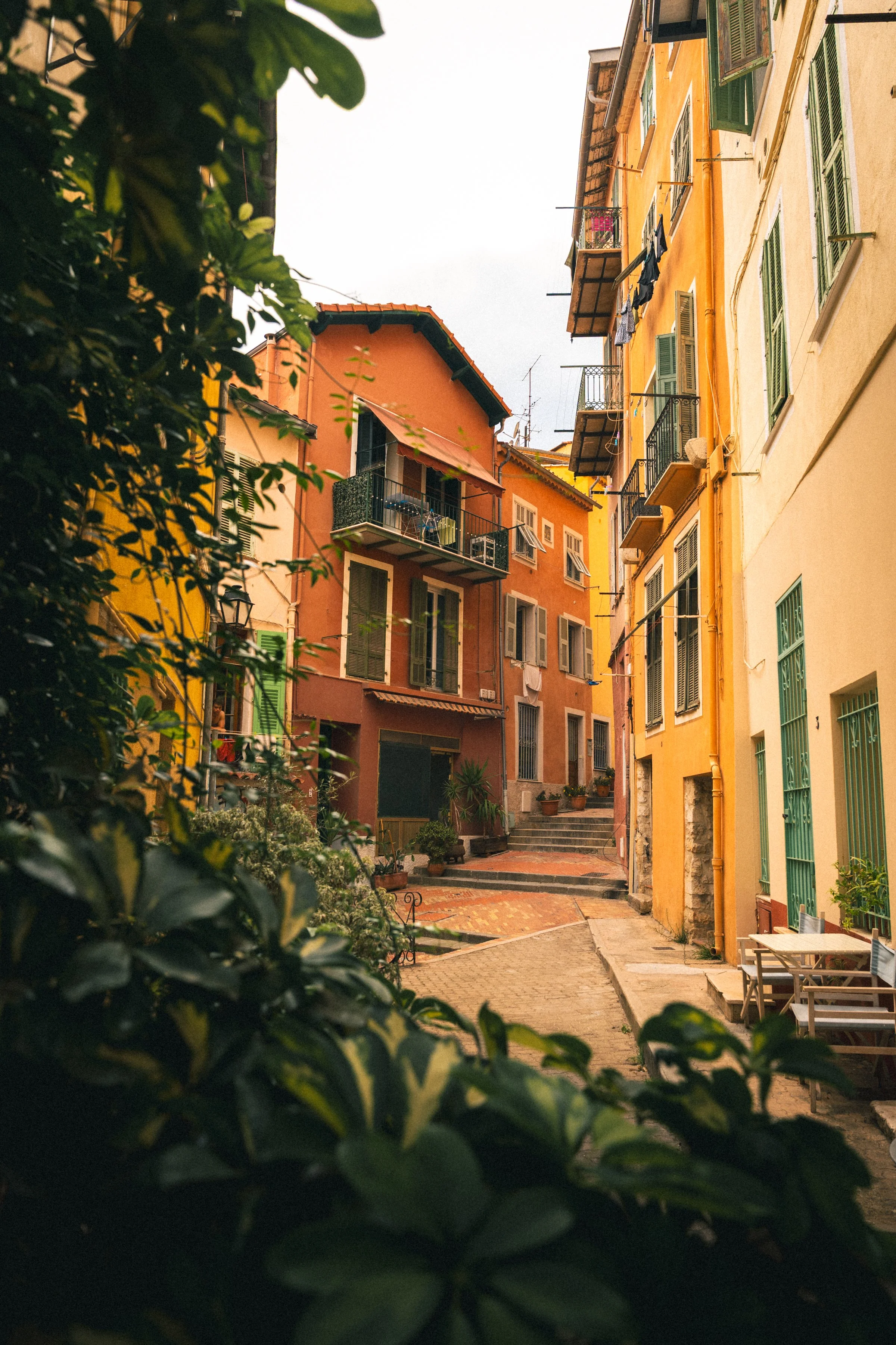 Colorful European street with yellow and orange buildings, green shutters, balconies, potted plants, and outdoor seating under an overcast sky.