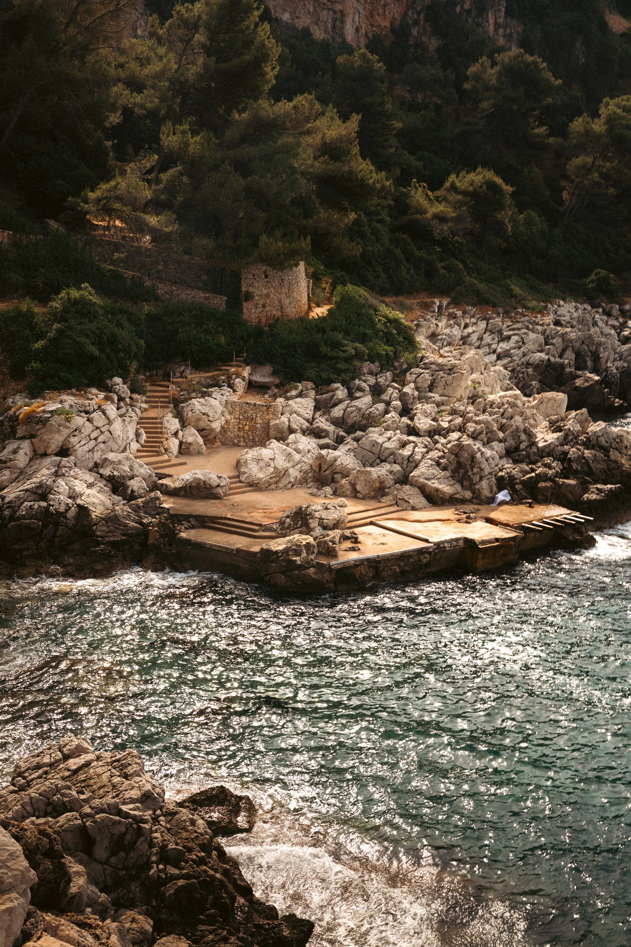 Rocky coastline with stairs leading down to the water, surrounded by green trees and hills in the background.